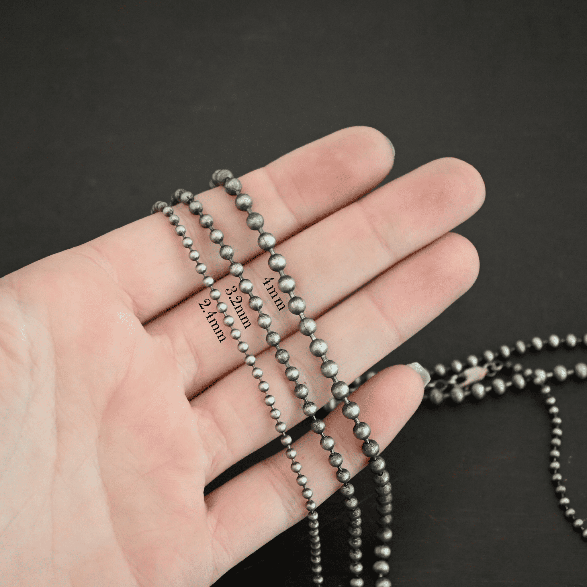 Beau Ball Chain Close up of the "Beau Ball" Chain with 4mm, 3.2 mm, and 2.4mm ball chains. Made out of brushed stainless steel with balls that are spaced apart evenly on the chain. Chains are shown against the fingertips of a hand. The highest chain in the photo is the 4mm chain, with the 3.2mm chain in the middle, and the 2.4mm chain at the bottom of the photo.