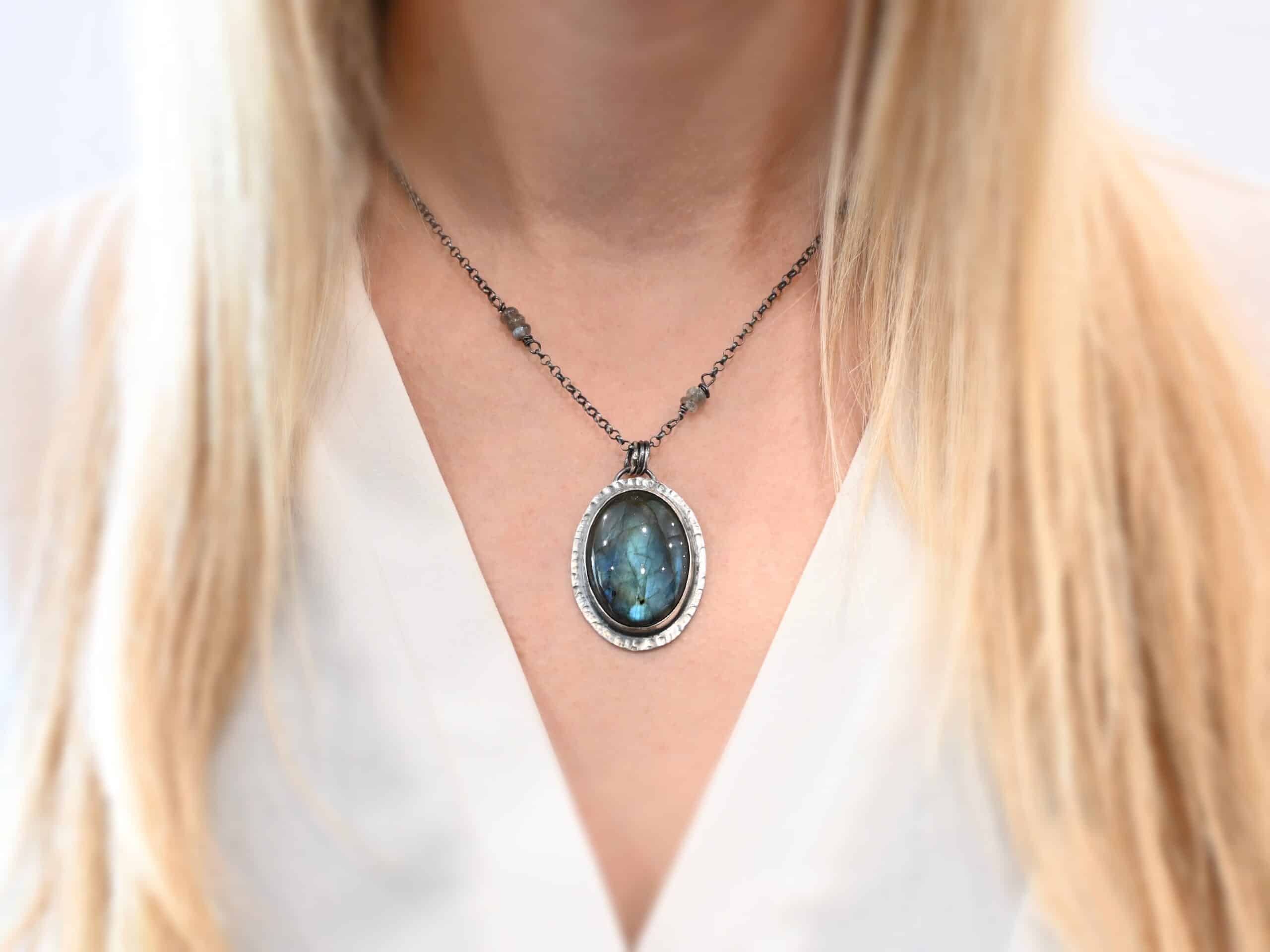 Raindrop Necklace Close up of the "Rain Drop" Necklace as worn on a model. This necklace is made out of sterling silver and labradorite with a sterling silver chain that has labradorite beads on the left and right side. The beads do not hang down from the chain. Rather, it looks as if it is the chain, then the beads, and then the chain continues. The large oval labradorite is the center stone and is set in sterling silver that is hammered around the edges, and then hung onto the chain with jump rings from a loop on the back of the pendant.