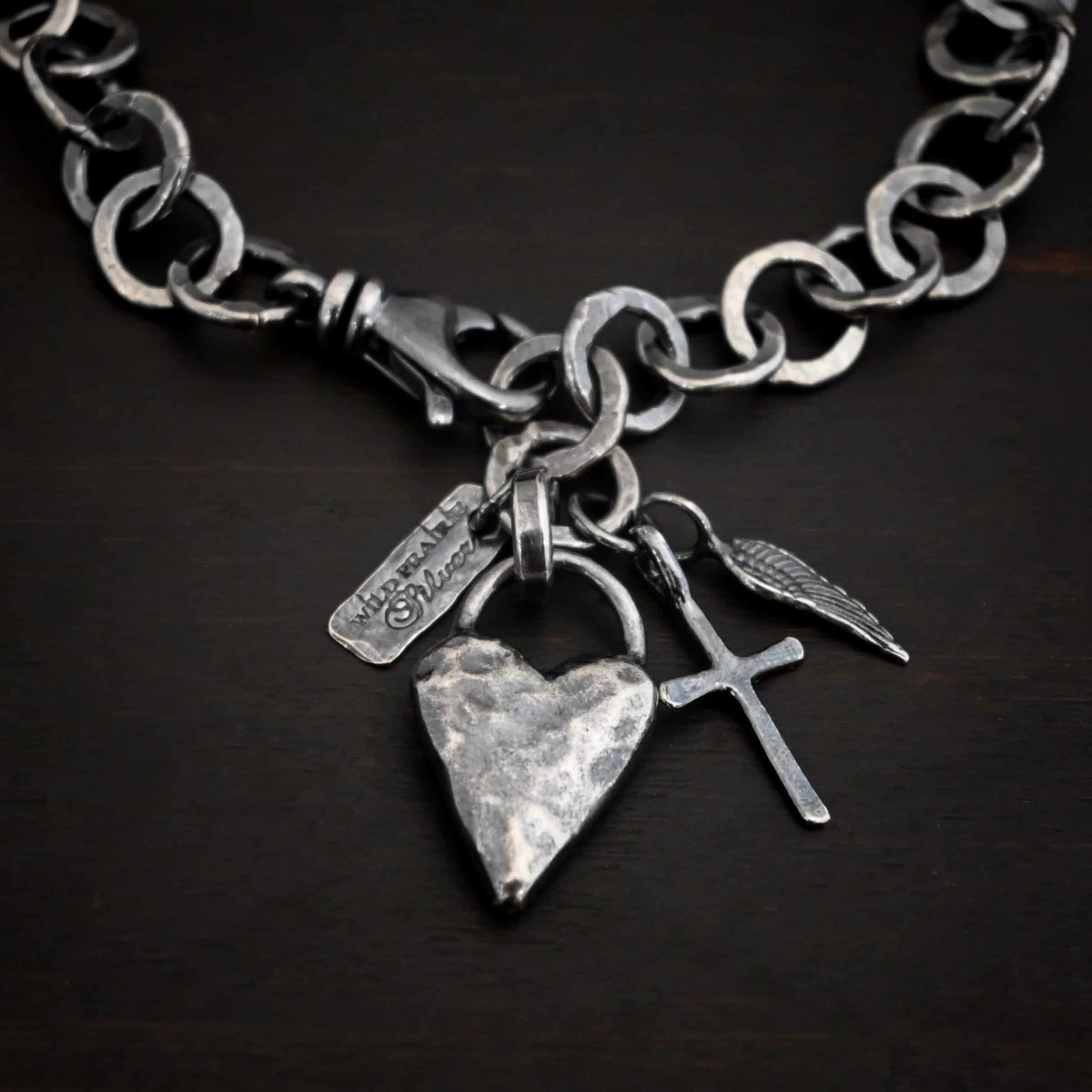 Mi Amour Bracelet Photo of the "Mi Amour" Bracelet. Bracelet has hammered and oxidized, thicker silver links for the chain of the bracelet. Connected to the end of the chain is a hammered, thick sterling silver heart that is oxidized, as well as a small sterling silver cross and angel wing charm, each with loops soldered to the top. There is also a small rectangle tag with a jump ring through it that has "Wild Prairie Silver" stamped into it. Bracelet is lying against a dark background.