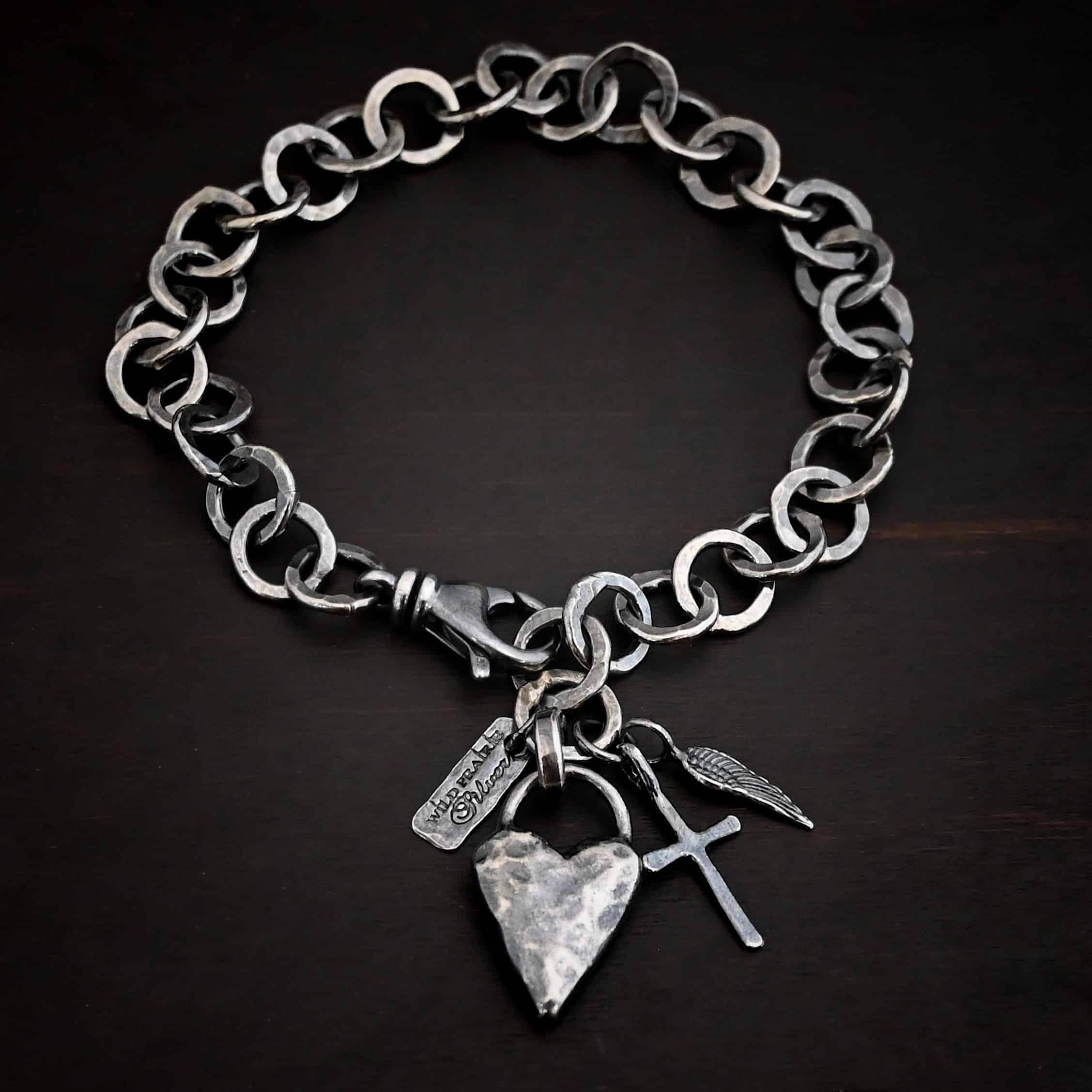 Mi Amour Bracelet Photo of the "Mi Amour" Bracelet. Bracelet has hammered and oxidized, thicker silver links for the chain of the bracelet. Connected to the end of the chain is a hammered, thick sterling silver heart that is oxidized, as well as a small sterling silver cross and angel wing charm, each with loops soldered to the top. There is also a small rectangle tag with a jump ring through it that has "Wild Prairie Silver" stamped into it. Bracelet is lying against a dark background.