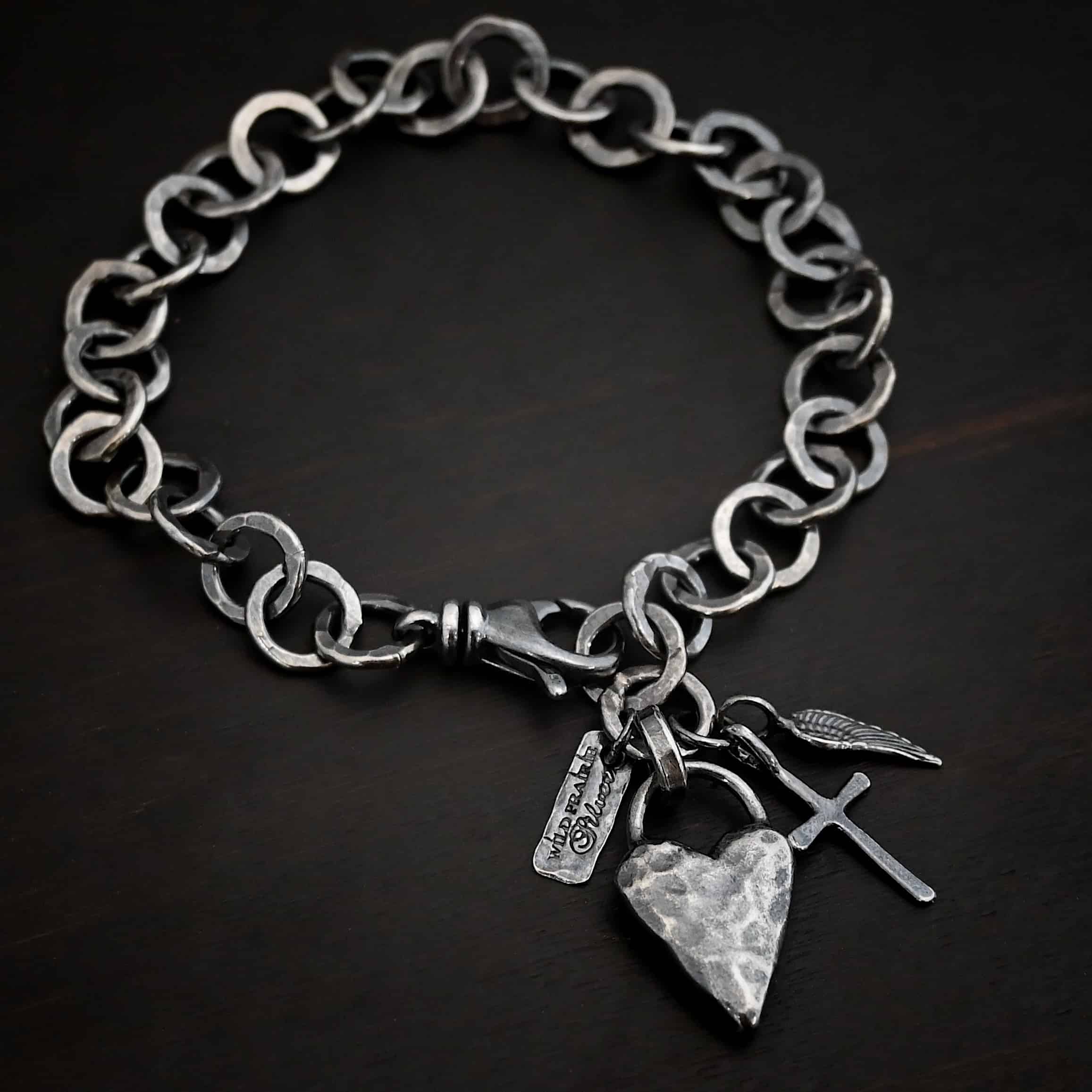 Mi Amour Bracelet Photo of the "Mi Amour" Bracelet. Bracelet has hammered and oxidized, thicker silver links for the chain of the bracelet. Connected to the end of the chain is a hammered, thick sterling silver heart that is oxidized, as well as a small sterling silver cross and angel wing charm, each with loops soldered to the top. There is also a small rectangle tag with a jump ring through it that has "Wild Prairie Silver" stamped into it. Bracelet is lying against a dark background.