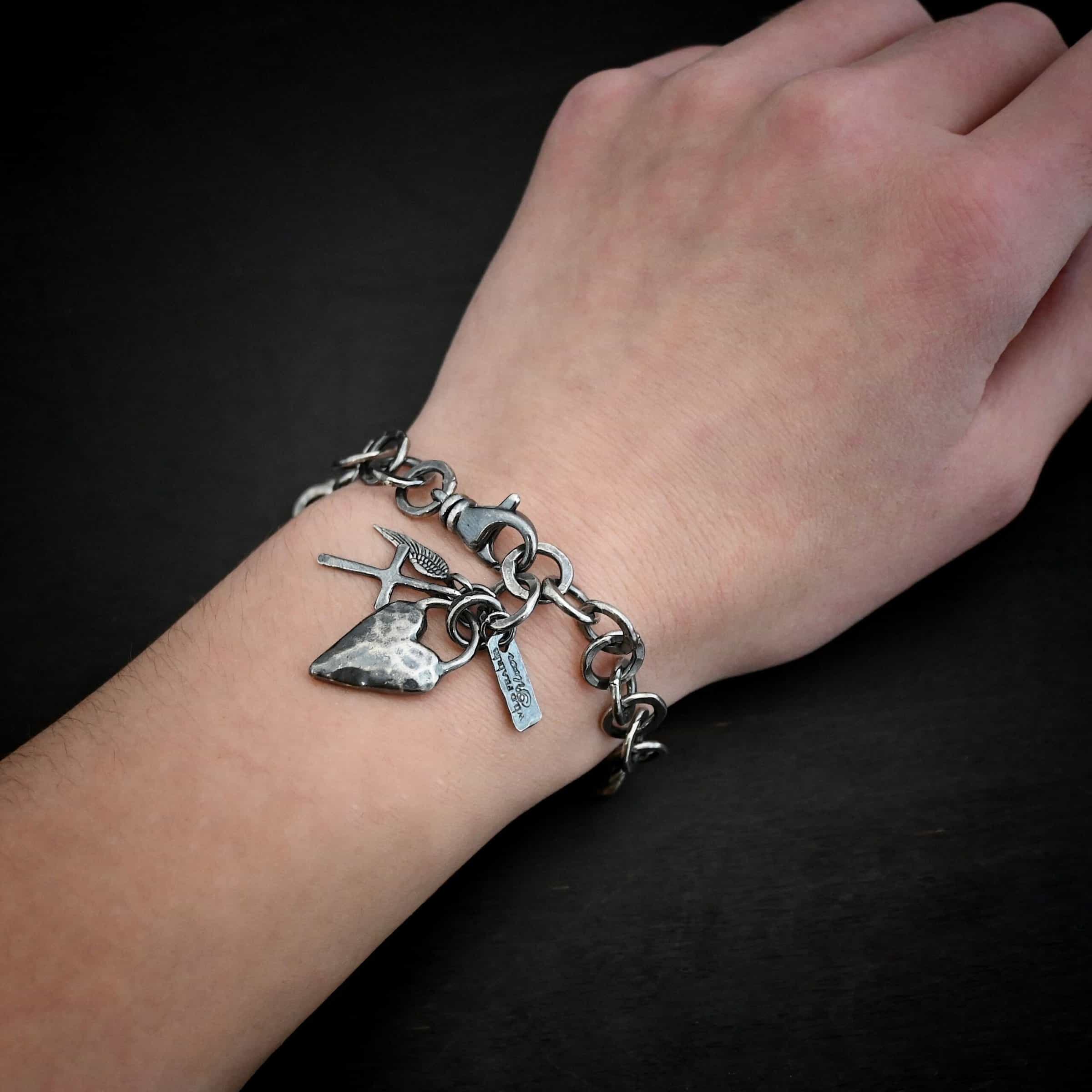 Mi Amour Bracelet Photo of the "Mi Amour" Bracelet. Bracelet has hammered and oxidized, thicker silver links for the chain of the bracelet. Connected to the end of the chain is a hammered, thick sterling silver heart that is oxidized, as well as a small sterling silver cross and angel wing charm, each with loops soldered to the top. There is also a small rectangle tag with a jump ring through it that has "Wild Prairie Silver" stamped into it. Bracelet is worn by a model up close.