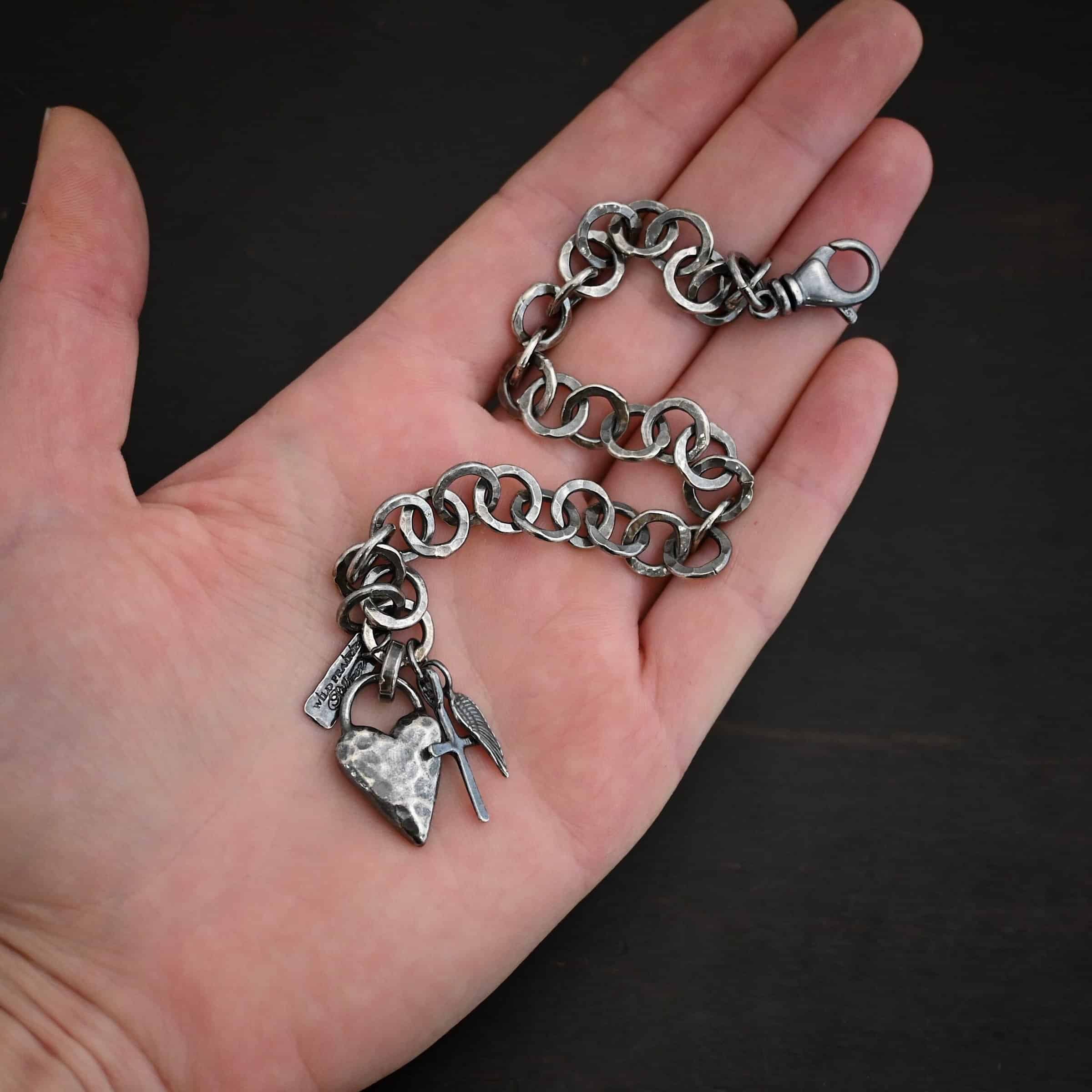 Mi Amour Bracelet Photo of the "Mi Amour" Bracelet. Bracelet has hammered and oxidized, thicker silver links for the chain of the bracelet. Connected to the end of the chain is a hammered, thick sterling silver heart that is oxidized, as well as a small sterling silver cross and angel wing charm, each with loops soldered to the top. There is also a small rectangle tag with a jump ring through it that has "Wild Prairie Silver" stamped into it. Bracelet is lying on the palm and fingertips of a hand.