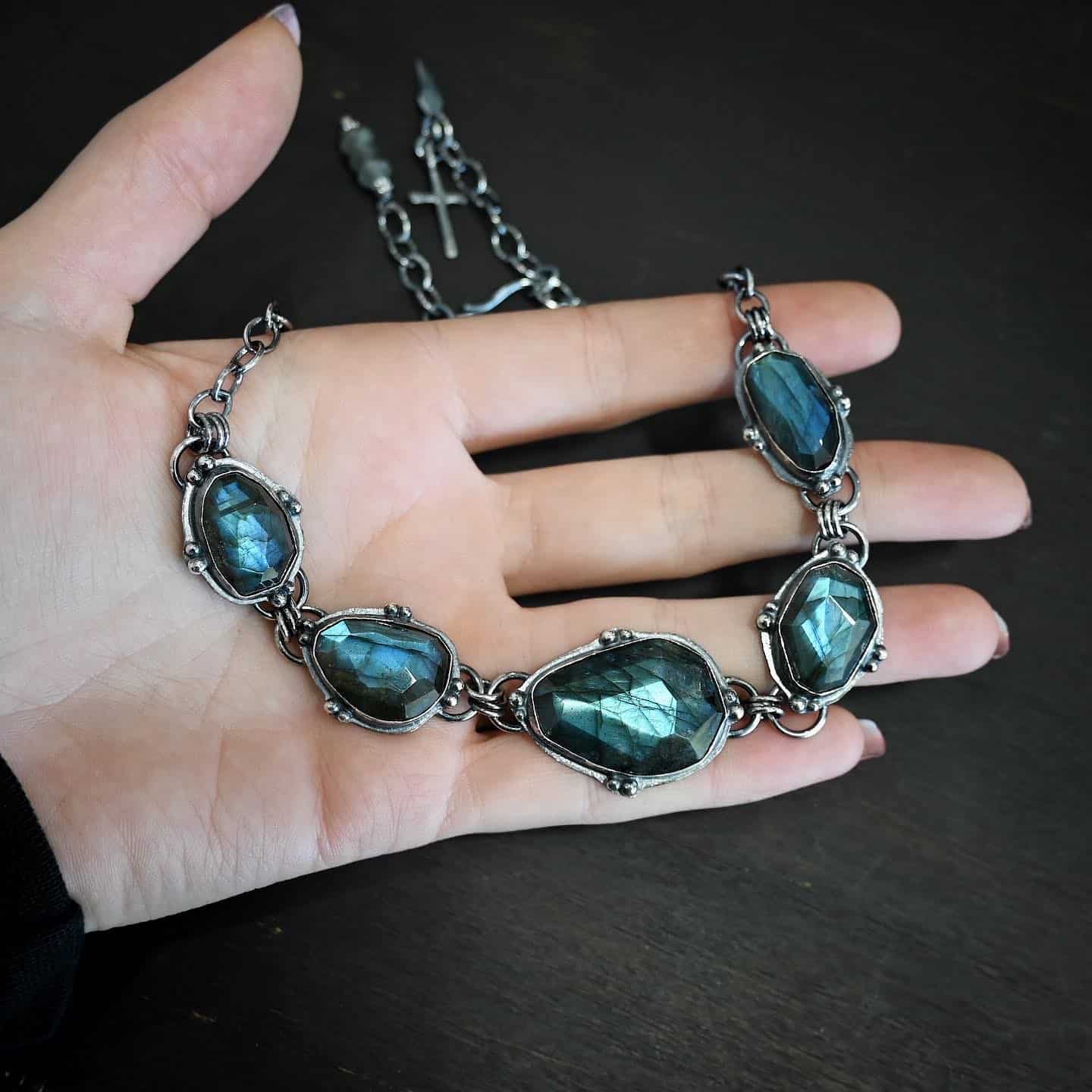 Ocean Whispers Necklace Photo of the "Ocean Whispers" Necklace. Made out of sterling silver and labradorite. There are five rose cut labradorite pieces, each with loops on the back and jump rings on the loops to attach each stone to one another. There are balls of silver scattered around each stone. On the two outermost labradorite stones, the chain of the necklace is attached to the loops, and there is a figure eight clasp at the back. Necklace is lying against the palm of a hand.