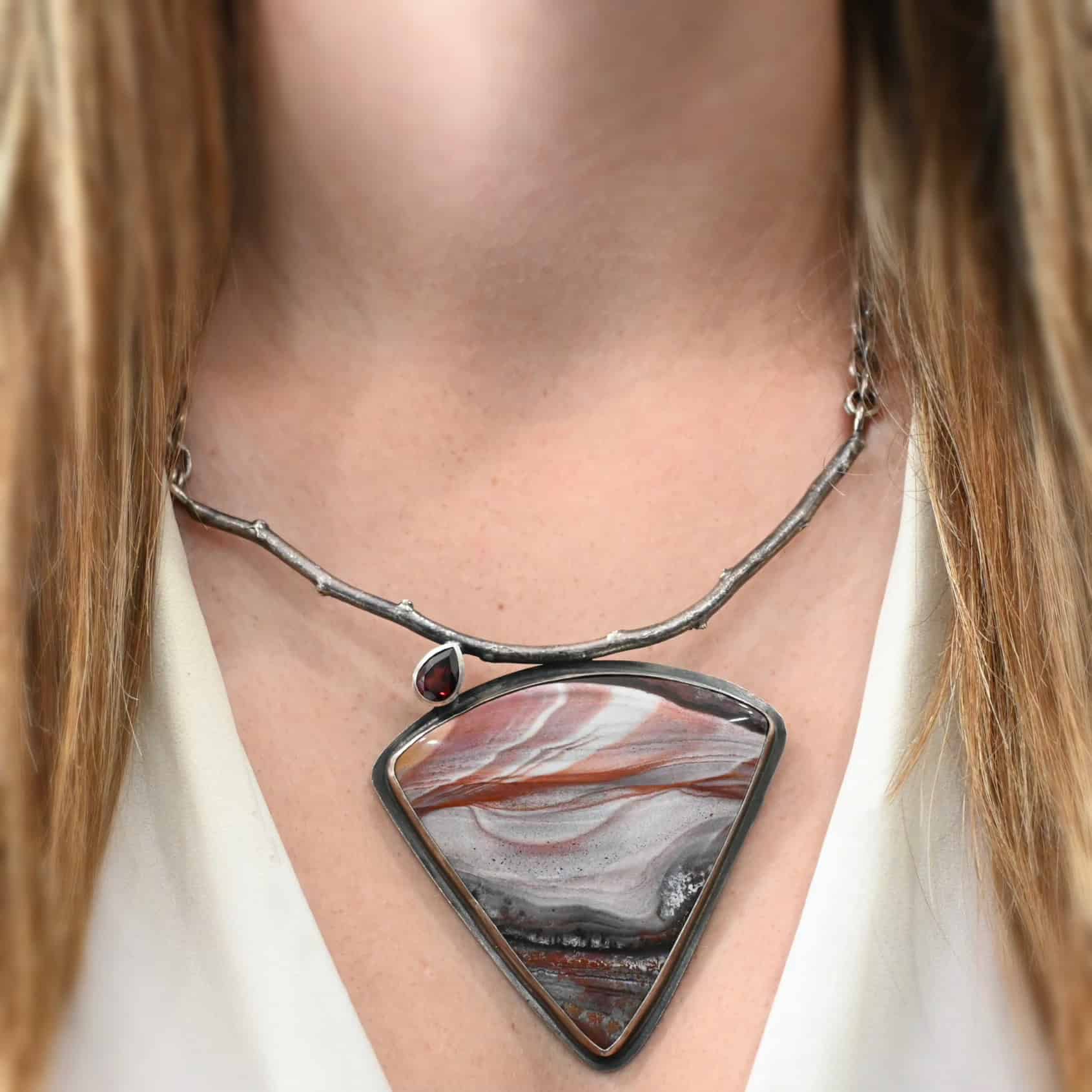 Borrowed From Someone Beautiful "Borrowed From Someone Beautiful" Necklace as shown on a model. Necklace is made out of landscape jasper and garnet. The landscape jasper is the large stone that is triangular shaped in the center. There is a teardrop shaped garnet in the left corner of the necklace. The jasper and garnet are both soldered and attached to a casted sterling silver large twig. The twig is attached on both sides to sterling silver chain. Model is wearing a white V-neck.