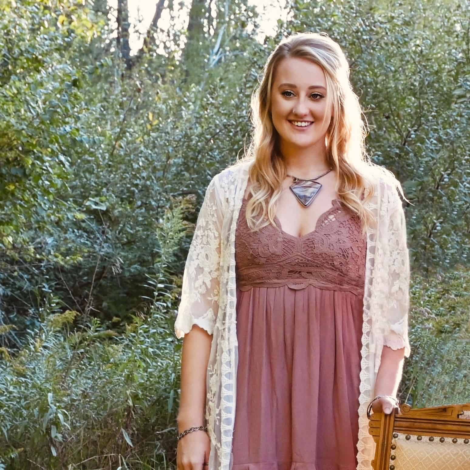 Borrowed from someone beautiful necklace Photo of Bekah. She is wearing a dress and a lace cardigan. Her hand is on a chair. She is wearing a large necklace. She is staring at the camera and smiling. She has long blonde hair that is curled.