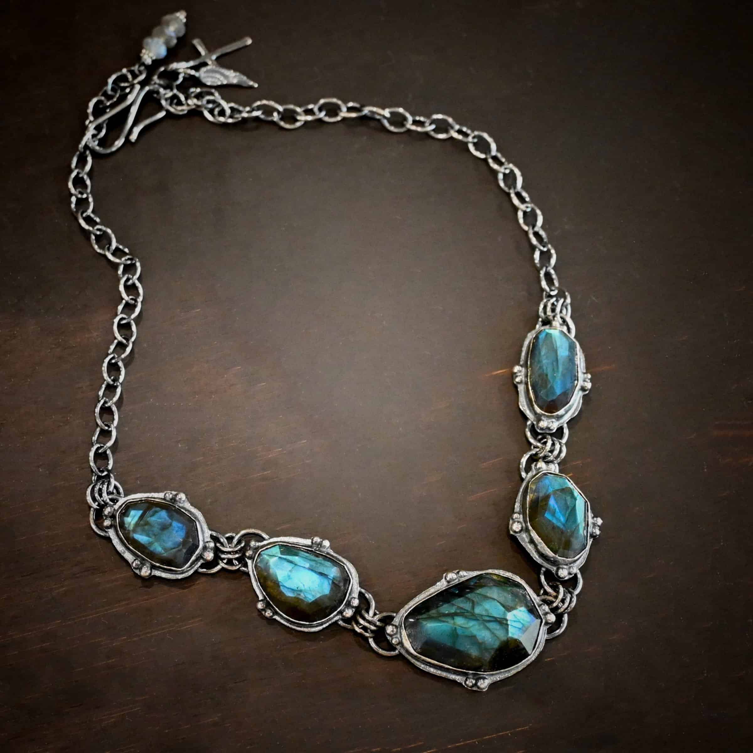 Ocean Whispers Necklace Photo of the "Ocean Whispers" Necklace. Made out of sterling silver and labradorite. There are five rose cut labradorite pieces, each with loops on the back and jump rings on the loops to attach each stone to one another. There are balls of silver scattered around each stone. On the two outermost labradorite stones, the chain of the necklace is attached to the loops, and there is a figure eight clasp at the back. Necklace is lying against a dark background.