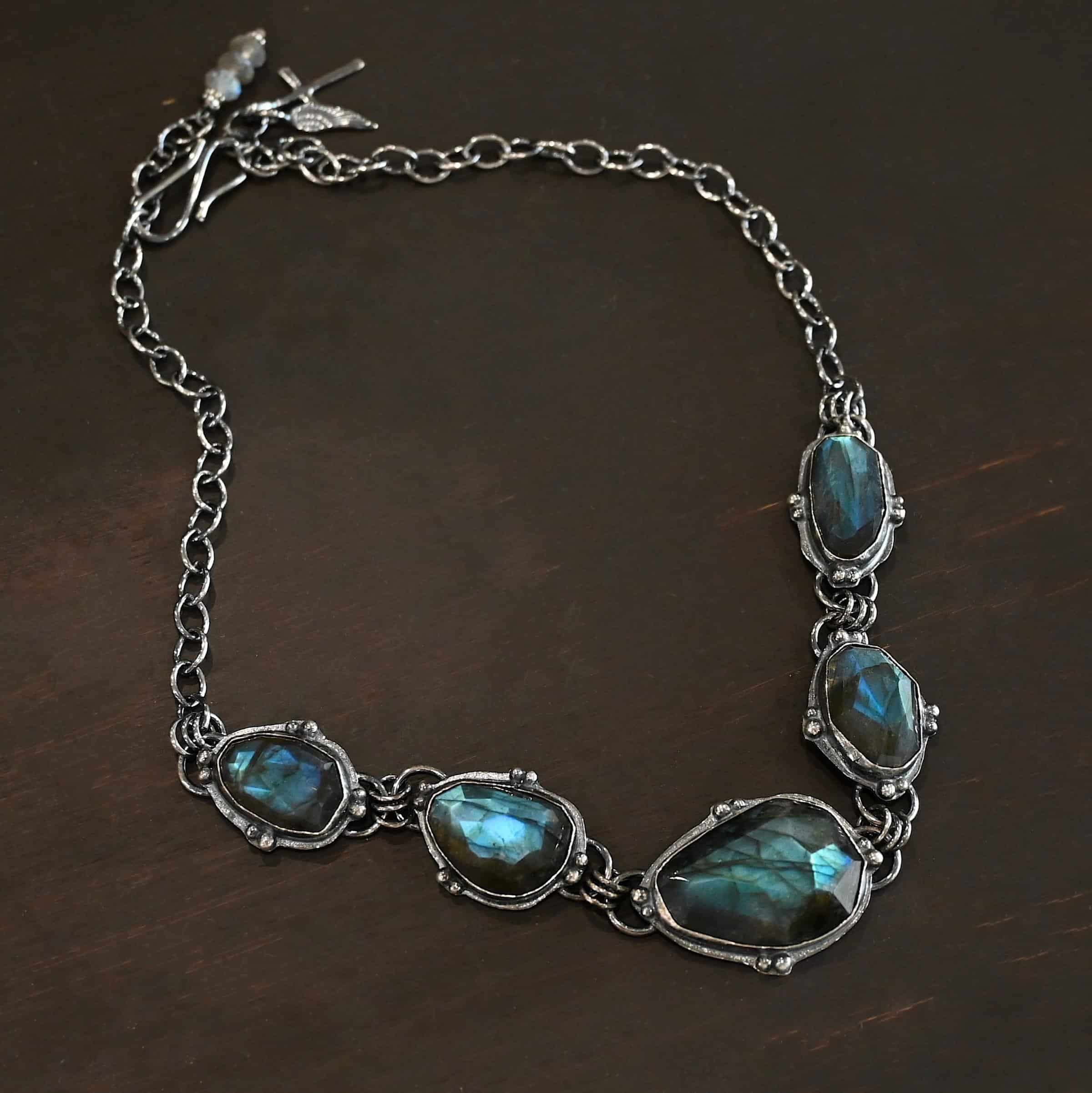 Ocean Whispers Necklace Photo of the "Ocean Whispers" Necklace. Made out of sterling silver and labradorite. There are five rose cut labradorite pieces, each with loops on the back and jump rings on the loops to attach each stone to one another. There are balls of silver scattered around each stone. On the two outermost labradorite stones, the chain of the necklace is attached to the loops, and there is a figure eight clasp at the back. Necklace is lying against a dark background.