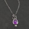 Close up of the "Bellflower" Necklace. Made out of sterling silver and amethyst. Amethyst is a slight oval, and is rose cut. Amethyst has three small silver dots on the top of it, and one small silver dot on the bottom, and is connected to the chain with three jump rings. Comes on a sterling silver "Cassandra" Chain. Necklace is set against a dark background.