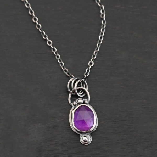 Close up of the "Bellflower" Necklace. Made out of sterling silver and amethyst. Amethyst is a slight oval, and is rose cut. Amethyst has three small silver dots on the top of it, and one small silver dot on the bottom, and is connected to the chain with three jump rings. Comes on a sterling silver "Cassandra" Chain. Necklace is set against a dark background.