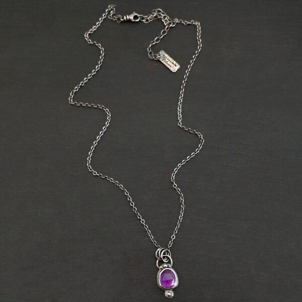 Close up of the "Bellflower" Necklace. Made out of sterling silver and amethyst. Amethyst is a slight oval, and is rose cut. Amethyst has three small silver dots on the top of it, and one small silver dot on the bottom, and is connected to the chain with three jump rings. Comes on a sterling silver "Cassandra" Chain. Necklace is set against a dark background.
