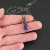 Close up of the "Bellflower" Necklace. Made out of sterling silver and amethyst. Amethyst is a slight oval, and is rose cut. Amethyst has three small silver dots on the top of it, and one small silver dot on the bottom, and is connected to the chain with three jump rings. Comes on a sterling silver "Cassandra" Chain. Necklace is set in the palm of a hand to demonstrate size. Hand is set against a dark background.