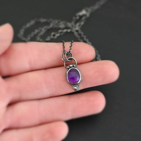 Close up of the "Bellflower" Necklace. Made out of sterling silver and amethyst. Amethyst is a slight oval, and is rose cut. Amethyst has three small silver dots on the top of it, and one small silver dot on the bottom, and is connected to the chain with three jump rings. Comes on a sterling silver "Cassandra" Chain. Necklace is set in the palm of a hand to demonstrate size. Hand is set against a dark background.