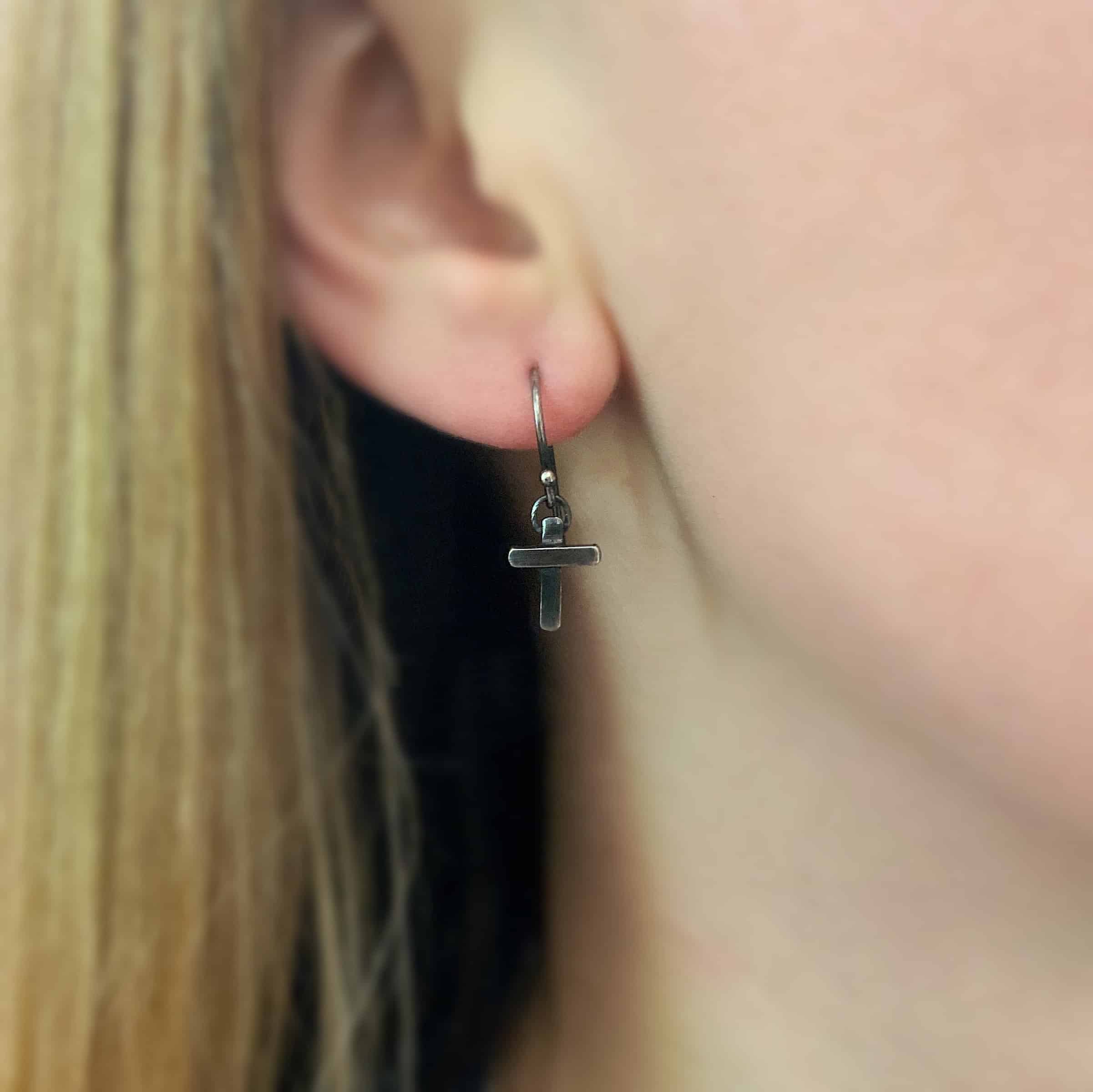 Photo of the "Old Rugged Cross" Earring as shown on a model. Made out of sterling silver and oxidized. Earring has a french hook ear wire with a jump ring on the bottom of it. Soldered to the jump ring is a small handmade cross charm. The cross has a small rectangle piece on the back, and a shorter rectangle piece soldered on top to the back piece. The shorter piece goes across the center of the back piece.