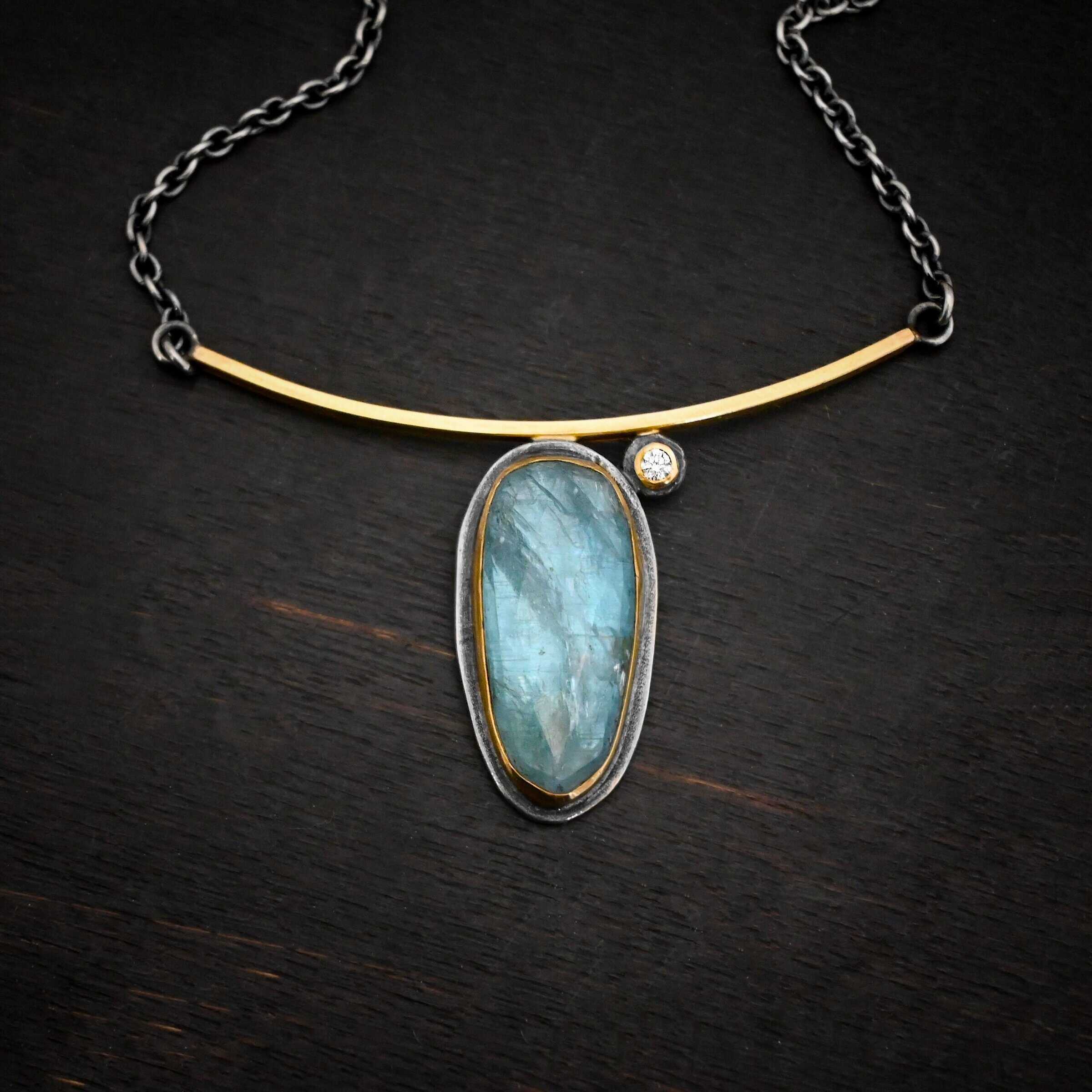 Photo of the "Day Dream" Necklace. Made out of sterling silver, gold, kyanite, and moissanite. There is an oval shaped kyanite set vertically in gold that is soldered to the bottom of a curved gold bar that is smooth. The kyanite has a silver back behind it to keep the stone in place. There is a small moissanite on the top right side of the kyanite. The moissanite is also soldered to the bar of the necklace. It is set in gold with silver behind it. The bar of gold has a silver loop on each end that each end of the chain attaches to. Necklace is lying against a dark background.