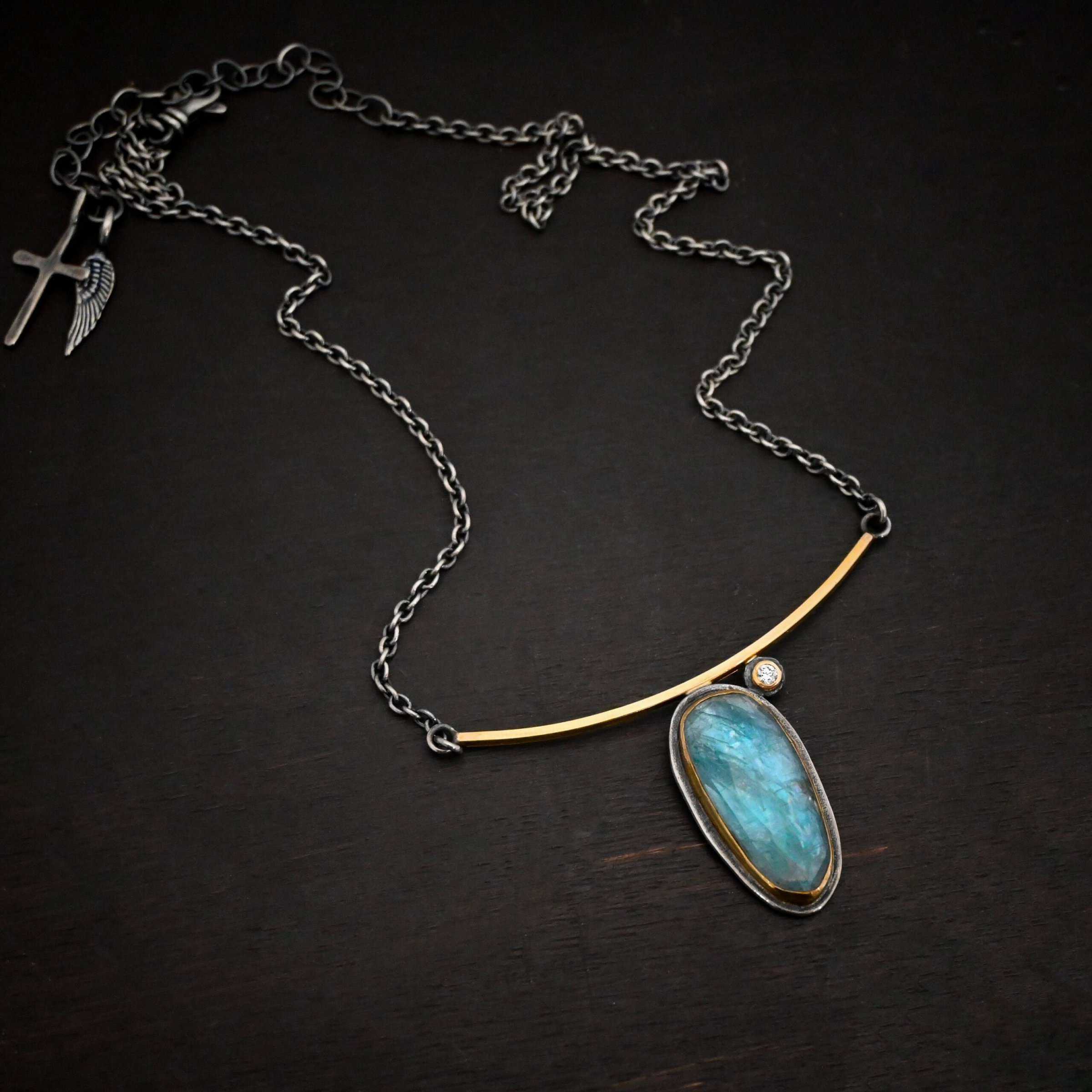 Photo of the "Day Dream" Necklace. Made out of sterling silver, gold, kyanite, and moissanite. There is an oval shaped kyanite set vertically in gold that is soldered to the bottom of a curved gold bar that is smooth. The kyanite has a silver back behind it to keep the stone in place. There is a small moissanite on the top right side of the kyanite. The moissanite is also soldered to the bar of the necklace. It is set in gold with silver behind it. The bar of gold has a silver loop on each end that each end of the chain attaches to. Necklace is lying against a dark background.