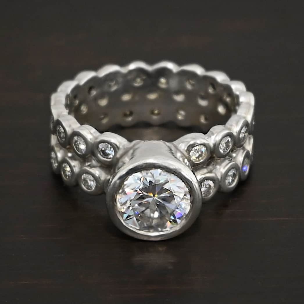 Heaven on Earth Ring Photo of the "Heaven On Earth" Ring. Made out of sterling silver and moissanites. There is a large moissanite in the center of the ring. The ring band is made of moissanites all the way around. There are two separate bands that are soldered together in the middle to create one band. Each band is made entirely out of moissanites that are set in silver and soldered together. Ring is sitting against a dark background.