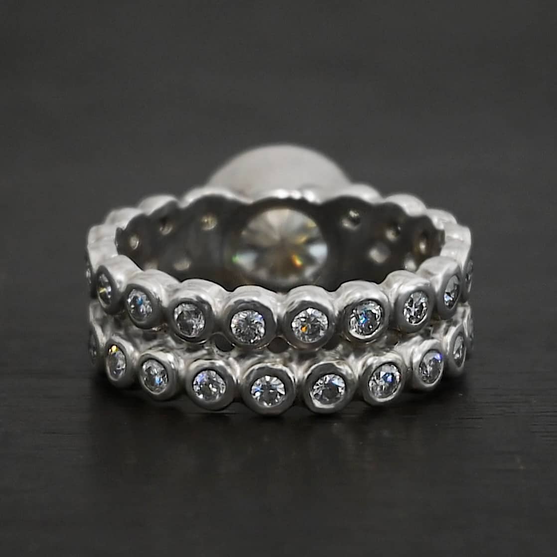 Heaven on Earth Ring Photo of the back of the "Heaven On Earth" Ring. Made out of sterling silver and moissanites. There is a large moissanite in the center of the ring. The ring band is made of moissanites all the way around. There are two separate bands that are soldered together in the middle to create one band. Each band is made entirely out of moissanites that are set in silver and soldered together. Ring is sitting against a dark background.