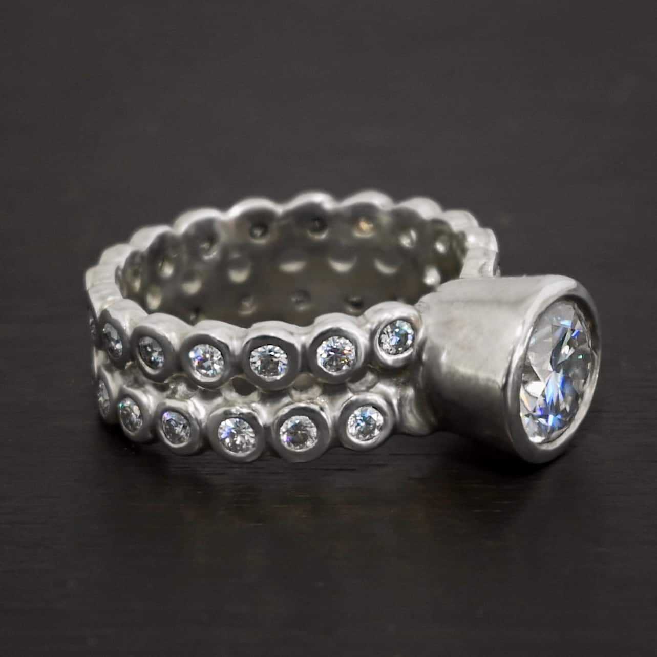 Heaven on Earth Ring Photo of the "Heaven On Earth" Ring. Made out of sterling silver and moissanites. There is a large moissanite in the center of the ring. The ring band is made of moissanites all the way around. There are two separate bands that are soldered together in the middle to create one band. Each band is made entirely out of moissanites that are set in silver and soldered together. Ring is sitting against a dark background.