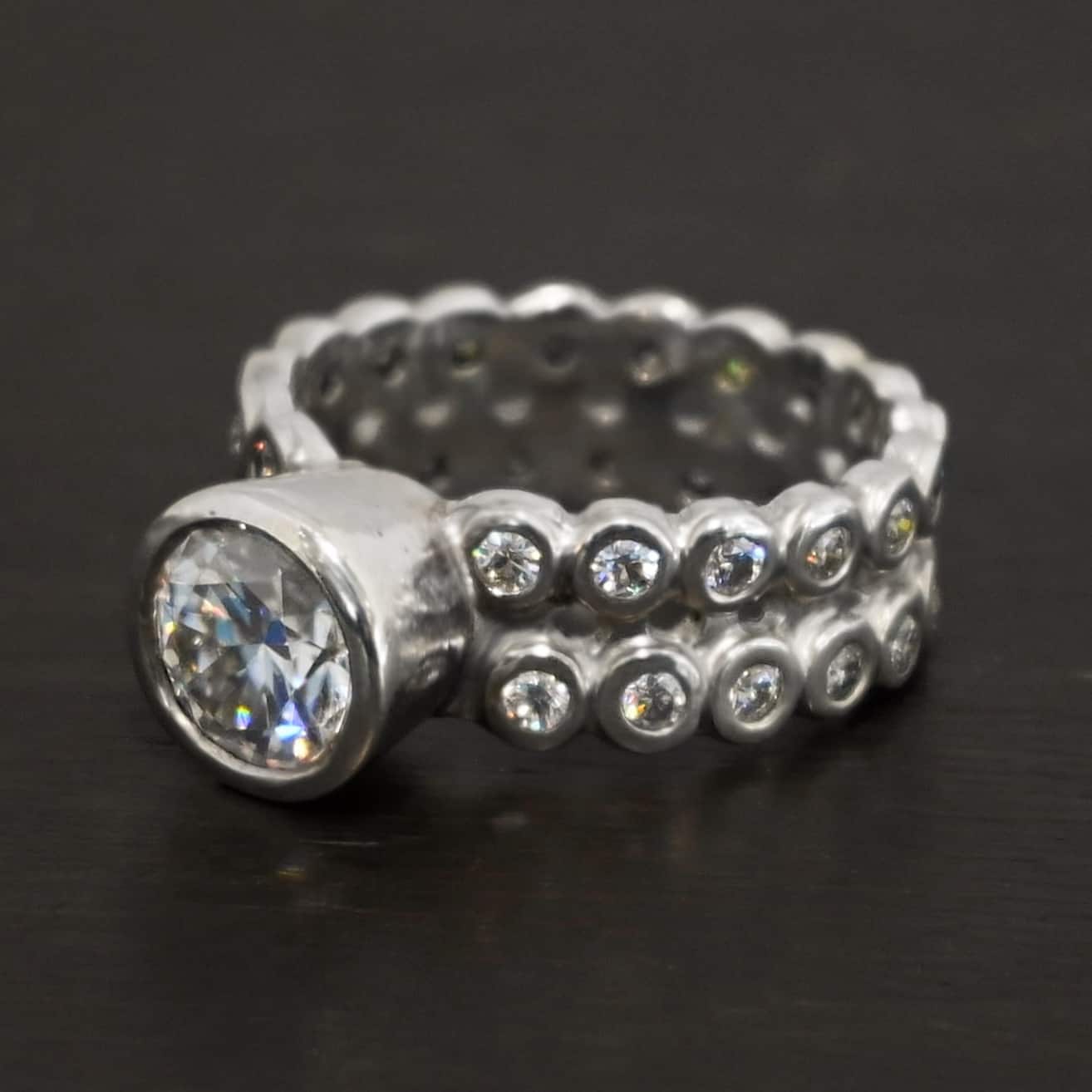 Heaven on Earth Ring Photo of the "Heaven On Earth" Ring. Made out of sterling silver and moissanites. There is a large moissanite in the center of the ring. The ring band is made of moissanites all the way around. There are two separate bands that are soldered together in the middle to create one band. Each band is made entirely out of moissanites that are set in silver and soldered together. Ring is sitting against a dark background.