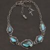Photo of the "Ocean Whispers" Necklace. Made out of sterling silver and labradorite. There are five rose cut labradorite pieces, each with loops on the back and jump rings on the loops to attach each stone to one another. There are balls of silver scattered around each stone. On the two outermost labradorite stones, the chain of the necklace is attached to the loops, and there is a figure eight clasp at the back. Necklace is lying against a dark background.