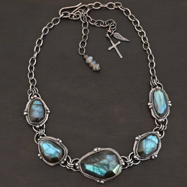 Photo of the "Ocean Whispers" Necklace. Made out of sterling silver and labradorite. There are five rose cut labradorite pieces, each with loops on the back and jump rings on the loops to attach each stone to one another. There are balls of silver scattered around each stone. On the two outermost labradorite stones, the chain of the necklace is attached to the loops, and there is a figure eight clasp at the back. Necklace is lying against a dark background.
