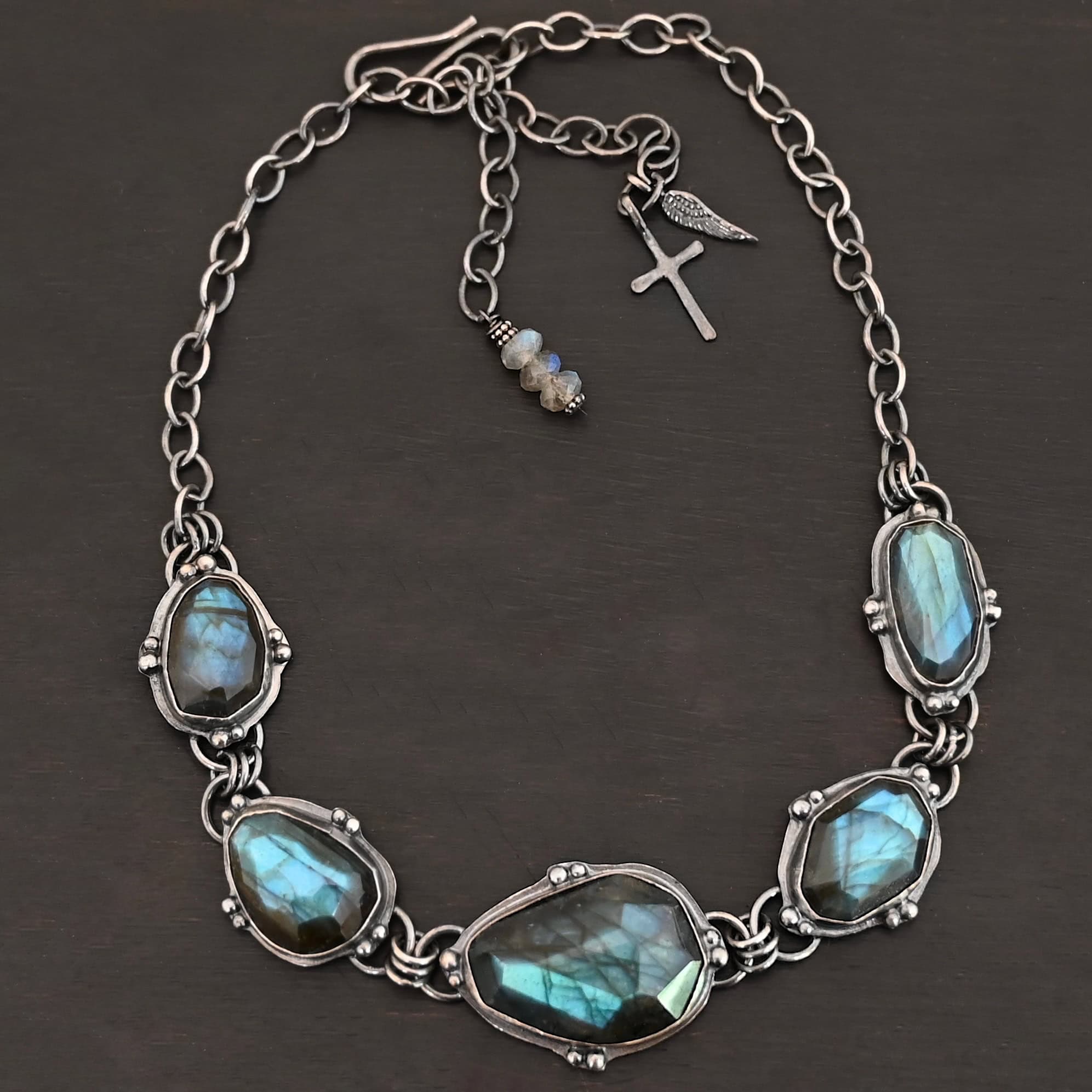 Ocean Whispers Necklace Photo of the "Ocean Whispers" Necklace. Made out of sterling silver and labradorite. There are five rose cut labradorite pieces, each with loops on the back and jump rings on the loops to attach each stone to one another. There are balls of silver scattered around each stone. On the two outermost labradorite stones, the chain of the necklace is attached to the loops, and there is a figure eight clasp at the back. Necklace is lying against a dark background.