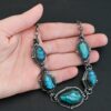 Photo of the "Ocean Whispers" Necklace. Made out of sterling silver and labradorite. There are five rose cut labradorite pieces, each with loops on the back and jump rings on the loops to attach each stone to one another. There are balls of silver scattered around each stone. On the two outermost labradorite stones, the chain of the necklace is attached to the loops, and there is a figure eight clasp at the back. Necklace is lying against the palm of a hand.