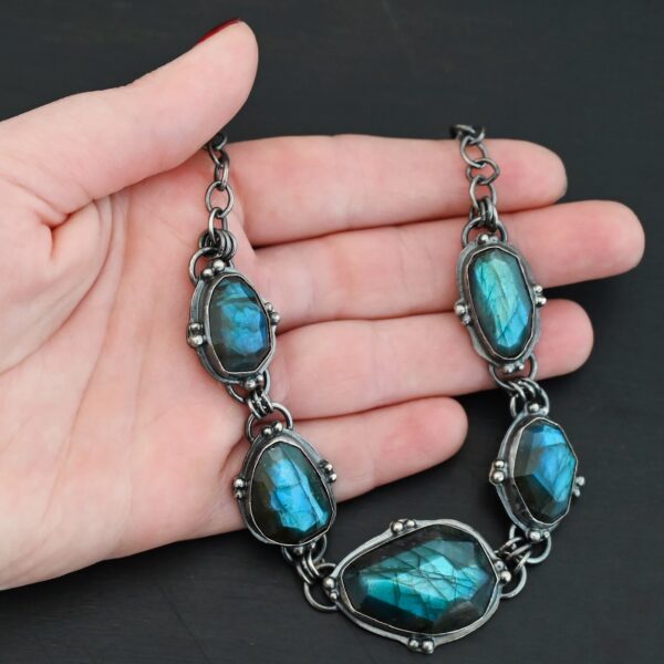 Photo of the "Ocean Whispers" Necklace. Made out of sterling silver and labradorite. There are five rose cut labradorite pieces, each with loops on the back and jump rings on the loops to attach each stone to one another. There are balls of silver scattered around each stone. On the two outermost labradorite stones, the chain of the necklace is attached to the loops, and there is a figure eight clasp at the back. Necklace is lying against the palm of a hand.