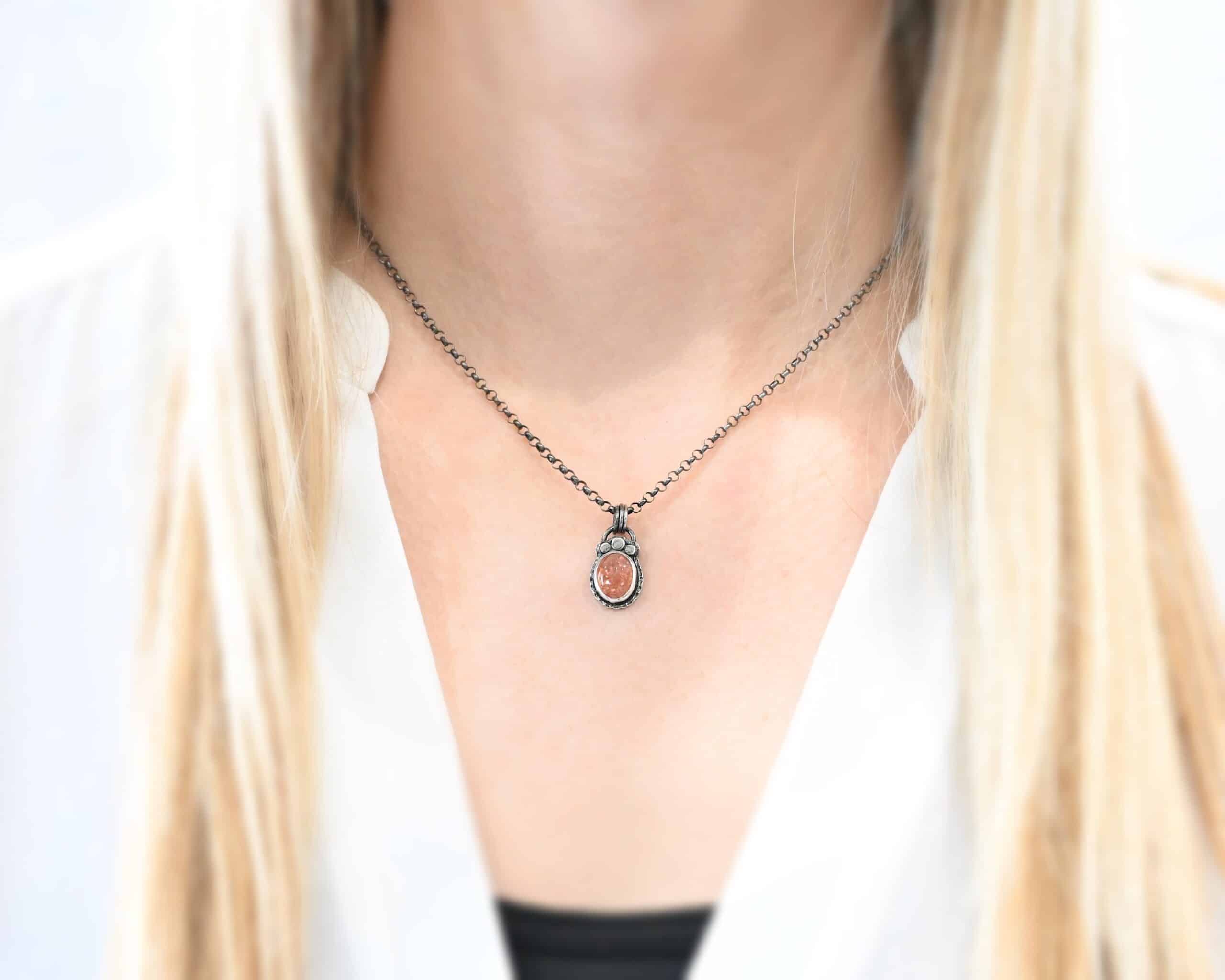 Photo of the "Ileah" Necklace. There is a small oval sunstone in the center of the pendant. There are three hammered silver dots on the top of it that follow the curve of the top of the oval. There is a loop soldered to the back with three silver jump rings going through it. The chain of the necklace goes through the jump rings. Necklace is worn by a model to demonstrate size.