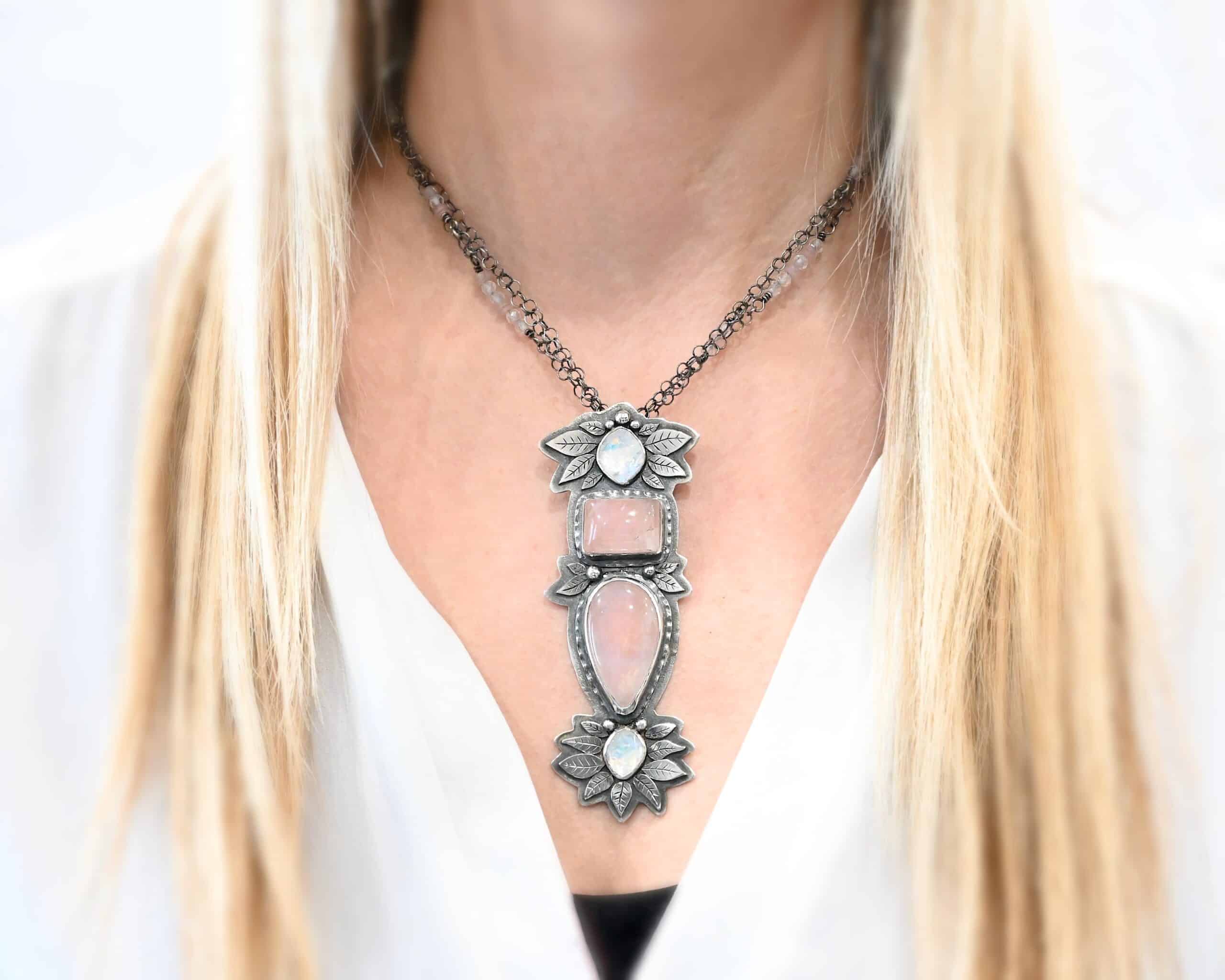 Sparkling Lady Necklace Photo of the "Sparkling Lady" Necklace. There is an oval moonstone at the top, a square rose quartz, an oval rose quartz, and another moonstone at the bottom. Surrounding the top, middle, and bottom are silver leaves that are engraved and balls of silver. Necklace is worn by a model to demonstrate size.
