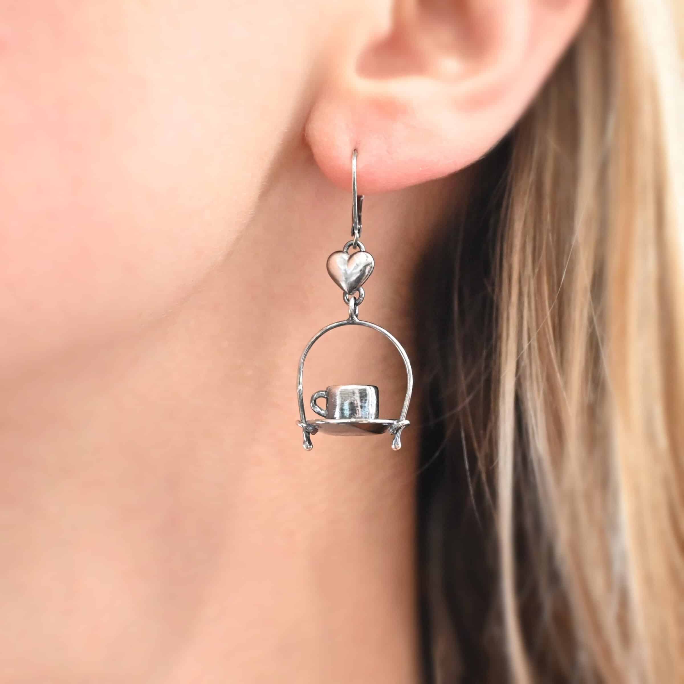 Photo of the "True Addiction" Coffee Earring shown up close by a model. Made out of sterling silver. Earring has an oxidized leverback ear wire. The bottom of each earwire has a sterling silver heart attached to it. Attached to the bottom of the heart is a large semicircle loop. In the center of the loop, attached on each side is a saucer and a coffee cup.