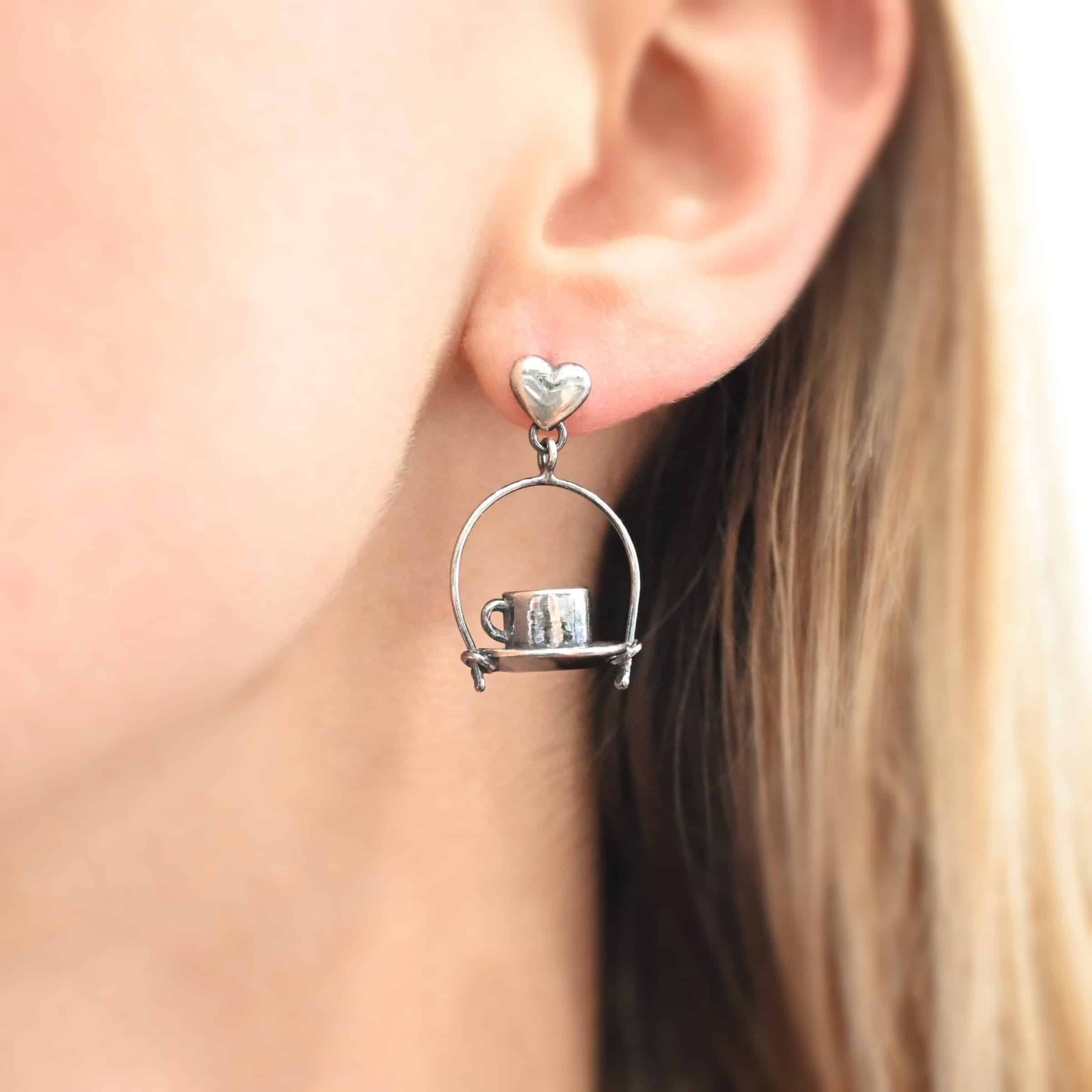 Photo of the "True Addiction" Coffee Earring as shown up close by a model. Made out of sterling silver. Earring has an oxidized post ear wire. The bottom of each earwire has a sterling silver heart attached to it. Attached to the bottom of the heart is a large semicircle loop. In the center of the loop, attached on each side is a saucer and a coffee cup.