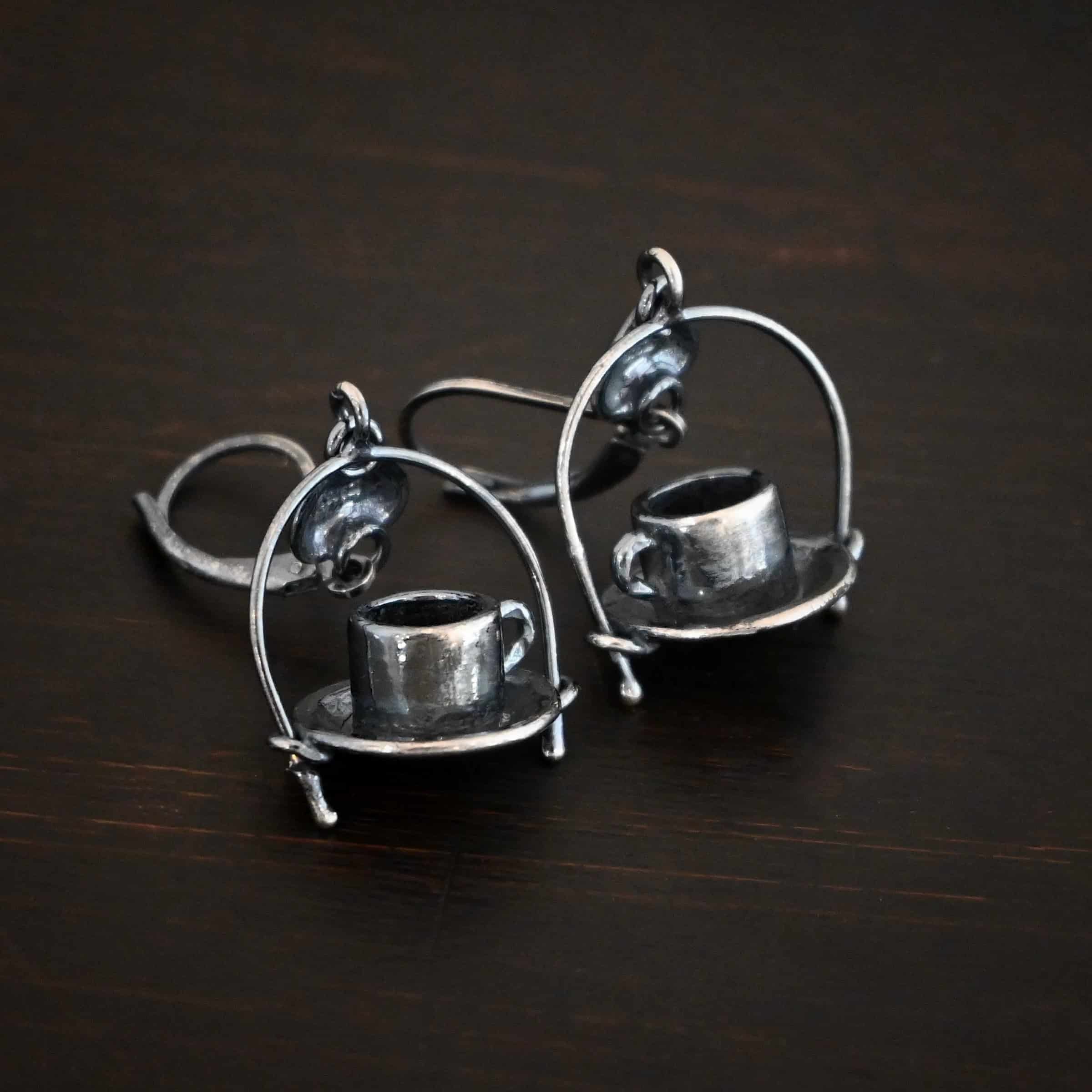 Photo of the "True Addiction" Coffee Earrings. Made out of sterling silver. Earrings have oxidized leverback ear wires. The bottom of each earwire has a sterling silver heart attached to it. Attached to the bottom of the heart is a large semicircle loop. In the center of the loop, attached on each side is a saucer and a coffee cup. Earrings are lying against a dark background.