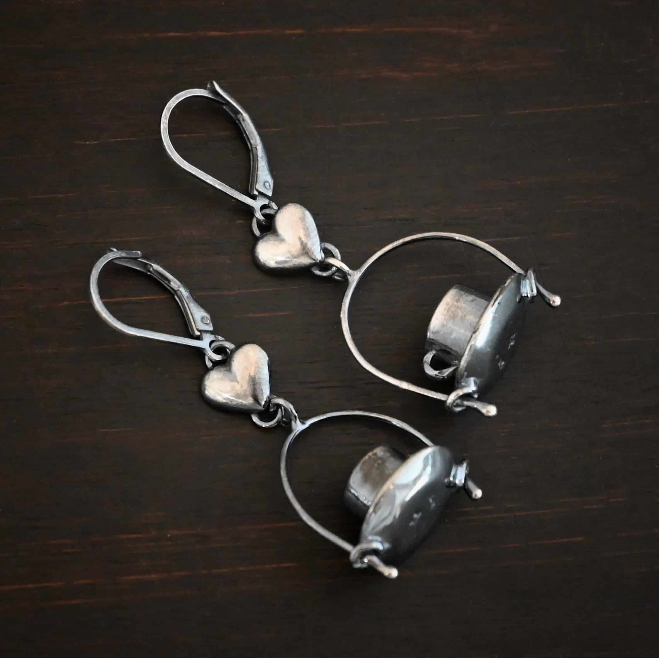 Photo of the "True Addiction" Coffee Earrings. Made out of sterling silver. Earrings have oxidized leverback ear wires. The bottom of each earwire has a sterling silver heart attached to it. Attached to the bottom of the heart is a large semicircle loop. In the center of the loop, attached on each side is a saucer and a coffee cup. Earrings are lying against a dark background.