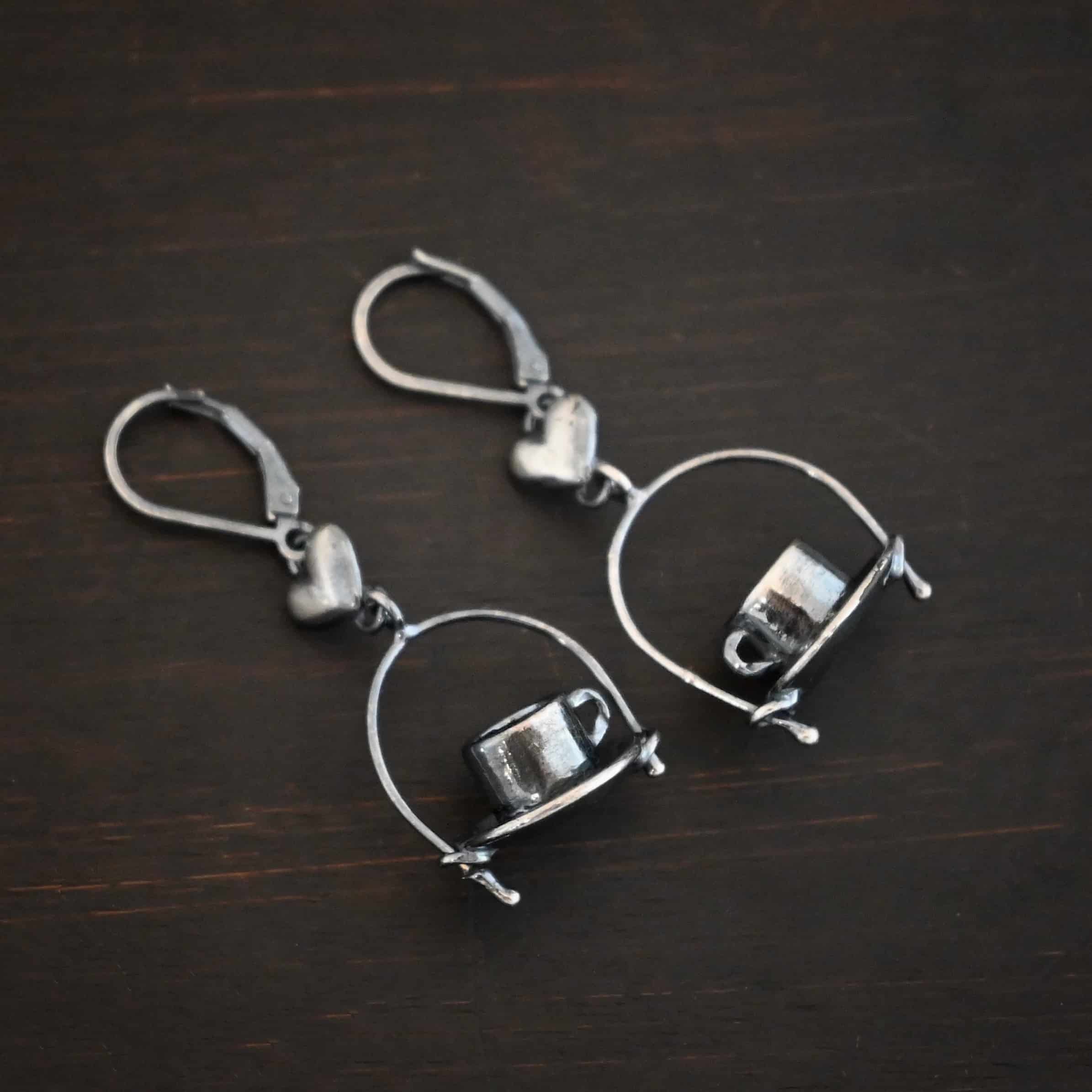 Photo of the "True Addiction" Coffee Earrings. Made out of sterling silver. Earrings have oxidized leverback ear wires. The bottom of each earwire has a sterling silver heart attached to it. Attached to the bottom of the heart is a large semicircle loop. In the center of the loop, attached on each side is a saucer and a coffee cup. Earrings are lying against a dark background.