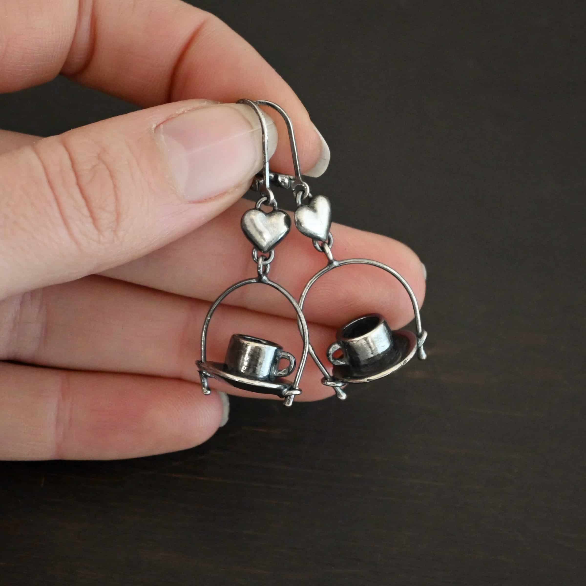 Photo of the "True Addiction" Coffee Earrings. Made out of sterling silver. Earrings have oxidized leverback ear wires. The bottom of each earwire has a sterling silver heart attached to it. Attached to the bottom of the heart is a large semicircle loop. In the center of the loop, attached on each side is a saucer and a coffee cup. Earrings are held in between the fingertips of a hand to demonstrate size.