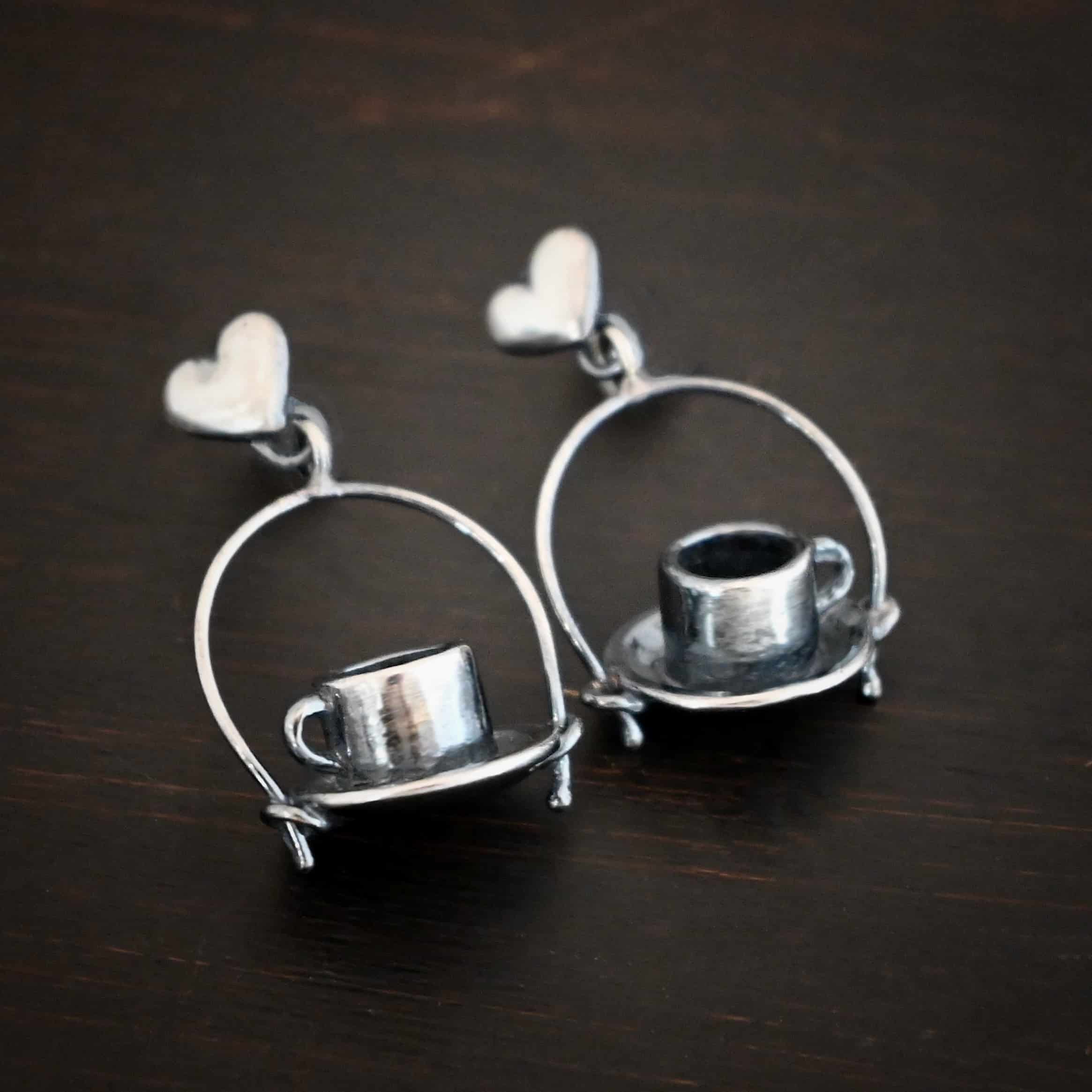 Photo of the "True Addiction" Coffee Earrings. Made out of sterling silver. Earrings have oxidized post ear wires. The bottom of each earwire has a sterling silver heart attached to it. Attached to the bottom of the heart is a large semicircle loop. In the center of the loop, attached on each side is a saucer and a coffee cup. Earrings are lying against a dark background.