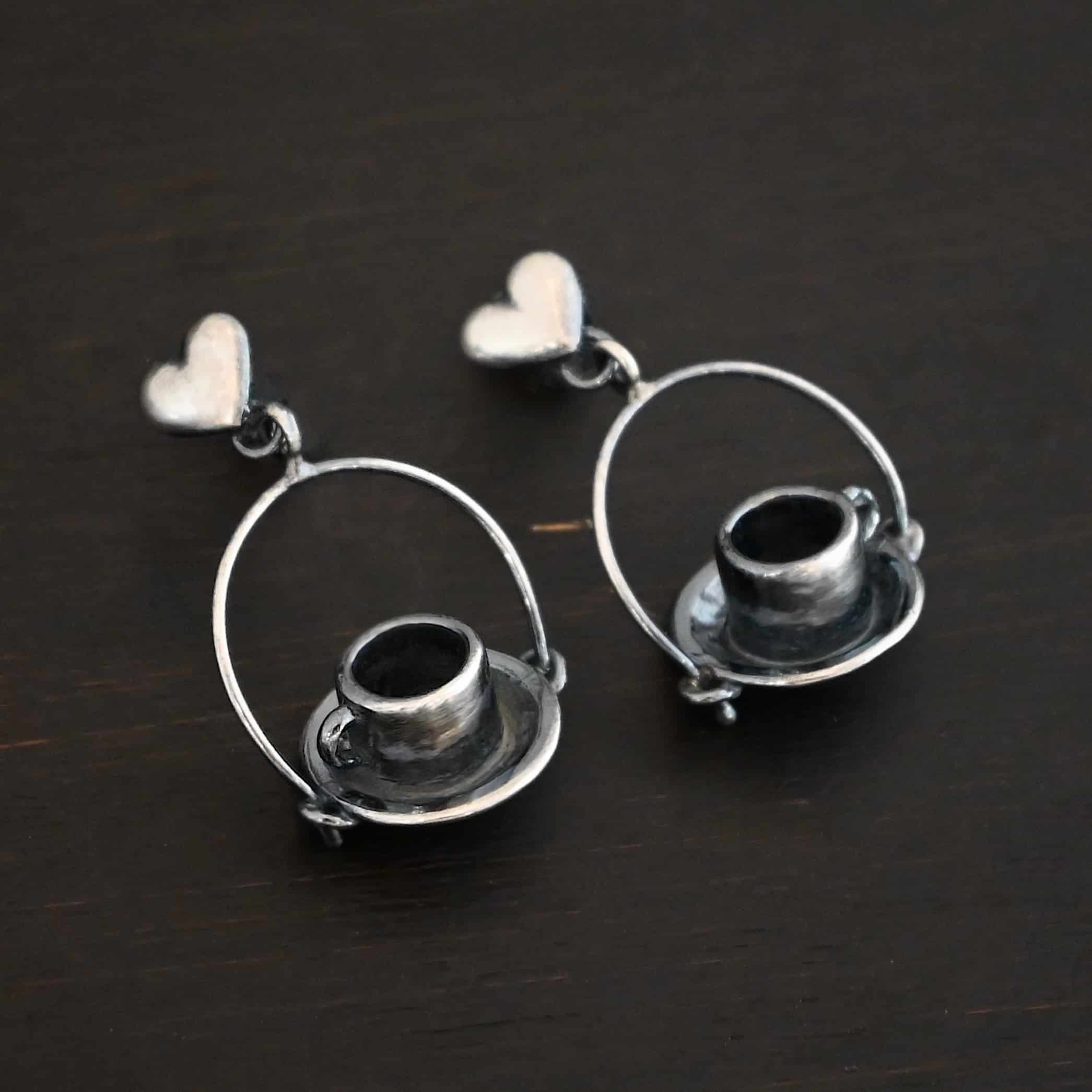 Photo of the "True Addiction" Coffee Earrings. Made out of sterling silver. Earrings have oxidized post ear wires. The bottom of each earwire has a sterling silver heart attached to it. Attached to the bottom of the heart is a large semicircle loop. In the center of the loop, attached on each side is a saucer and a coffee cup. Earrings are lying against a dark background.