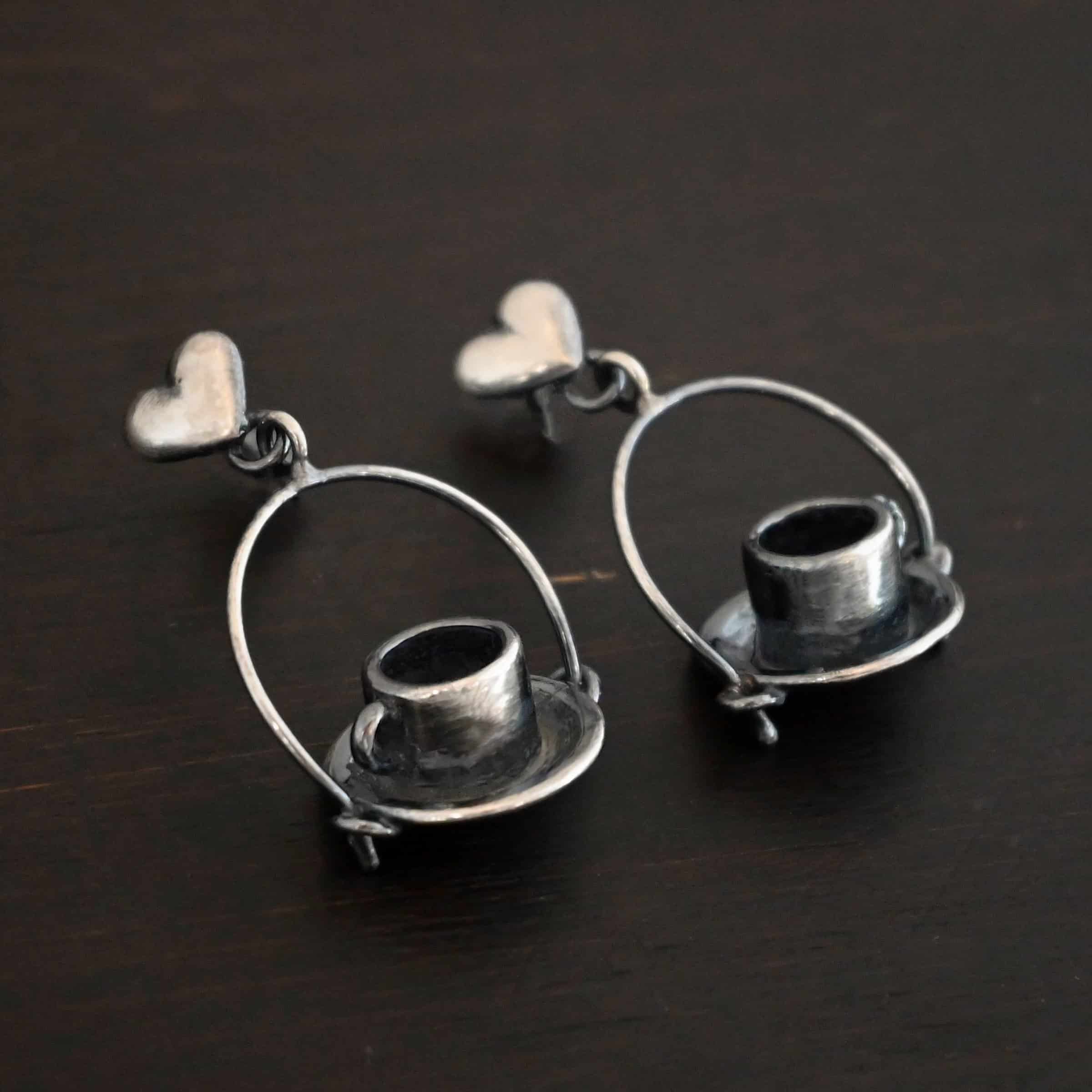 Photo of the "True Addiction" Coffee Earrings. Made out of sterling silver. Earrings have oxidized post ear wires. The bottom of each earwire has a sterling silver heart attached to it. Attached to the bottom of the heart is a large semicircle loop. In the center of the loop, attached on each side is a saucer and a coffee cup. Earrings are lying against a dark background.
