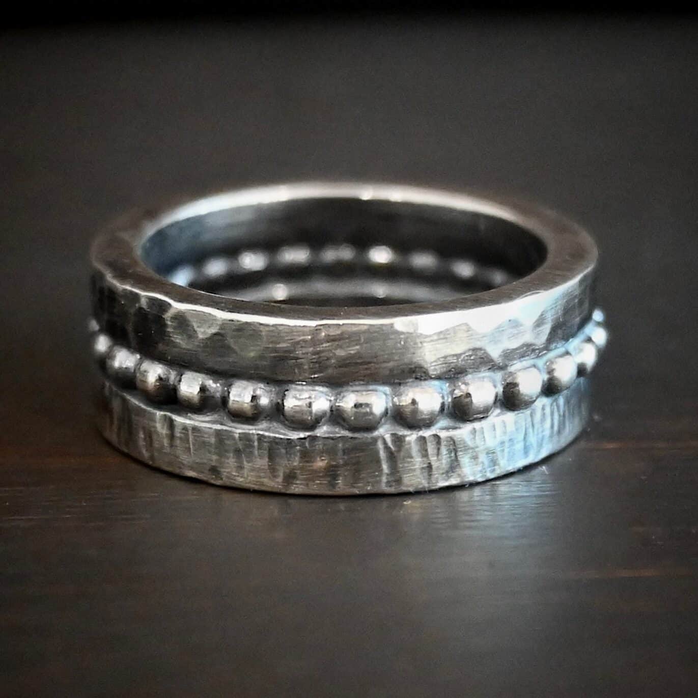 Trinity Ring Photo of the "Trinity" Ring. Made out of sterling silver. The top and bottom of the ring are hammered in different styles. The top is hammered with circles and the bottom has lines hammered into it. The middle of the ring has silver balls going all the way around it. The balls are set right next to one another all the way around the ring. Ring is sitting against a dark background.