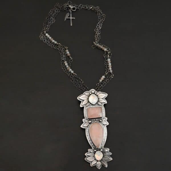 Close up of the "Sparkling Lady" Necklace. Made out of sterling silver, rose quartz, and moonstone. There are two rose quartz and two moonstone. The top of the pendant has an oval moonstone, with a square rose quartz beneath it. Beneath that is another rose quartz that is a teardrop shape with the point facing downwards. At the bottom of the pendant is another moonstone that is an oval shape. Surrounding each of these stones and around the entire pendant are silver leaves and dots. Pendant is laying against a dark background.