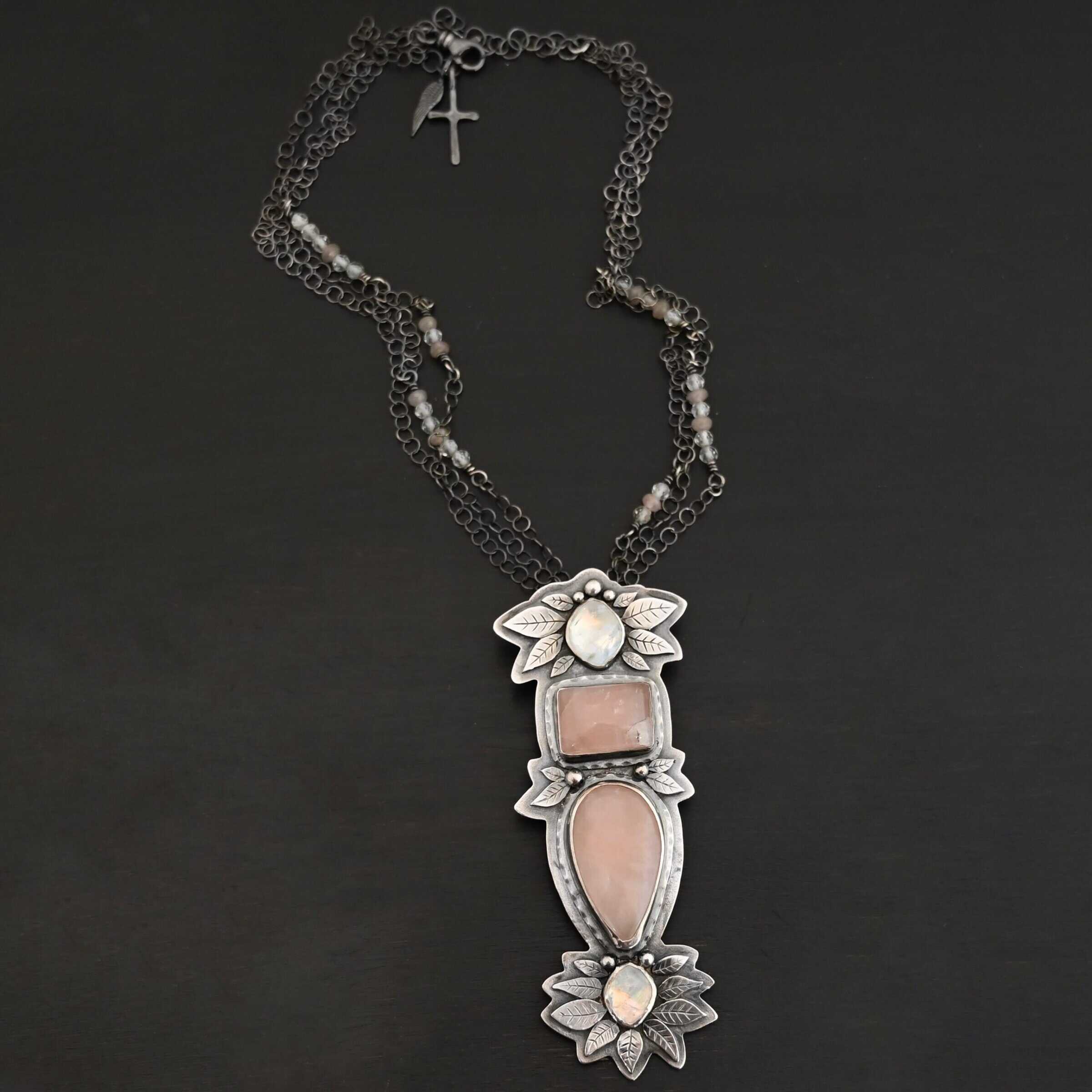 Sparkling Lady Necklace Close up of the "Sparkling Lady" Necklace. Made out of sterling silver, rose quartz, and moonstone. There are two rose quartz and two moonstone. The top of the pendant has an oval moonstone, with a square rose quartz beneath it. Beneath that is another rose quartz that is a teardrop shape with the point facing downwards. At the bottom of the pendant is another moonstone that is an oval shape. Surrounding each of these stones and around the entire pendant are silver leaves and dots. Pendant is laying against a dark background.