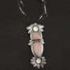Close up of the "Sparkling Lady" Necklace. Made out of sterling silver, rose quartz, and moonstone. There are two rose quartz and two moonstone. The top of the pendant has an oval moonstone, with a square rose quartz beneath it. Beneath that is another rose quartz that is a teardrop shape with the point facing downwards. At the bottom of the pendant is another moonstone that is an oval shape. Surrounding each of these stones and around the entire pendant are silver leaves and dots. Pendant is laying against a dark background.