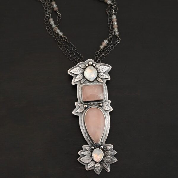 Close up of the "Sparkling Lady" Necklace. Made out of sterling silver, rose quartz, and moonstone. There are two rose quartz and two moonstone. The top of the pendant has an oval moonstone, with a square rose quartz beneath it. Beneath that is another rose quartz that is a teardrop shape with the point facing downwards. At the bottom of the pendant is another moonstone that is an oval shape. Surrounding each of these stones and around the entire pendant are silver leaves and dots. Pendant is laying against a dark background.