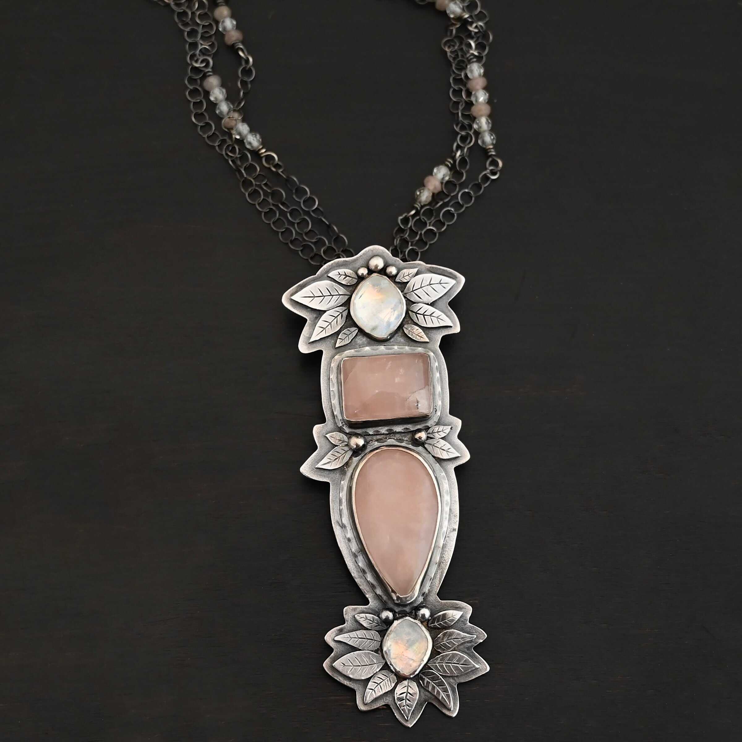 Sparkling Lady Necklace Close up of the "Sparkling Lady" Necklace. Made out of sterling silver, rose quartz, and moonstone. There are two rose quartz and two moonstone. The top of the pendant has an oval moonstone, with a square rose quartz beneath it. Beneath that is another rose quartz that is a teardrop shape with the point facing downwards. At the bottom of the pendant is another moonstone that is an oval shape. Surrounding each of these stones and around the entire pendant are silver leaves and dots. Pendant is laying against a dark background.