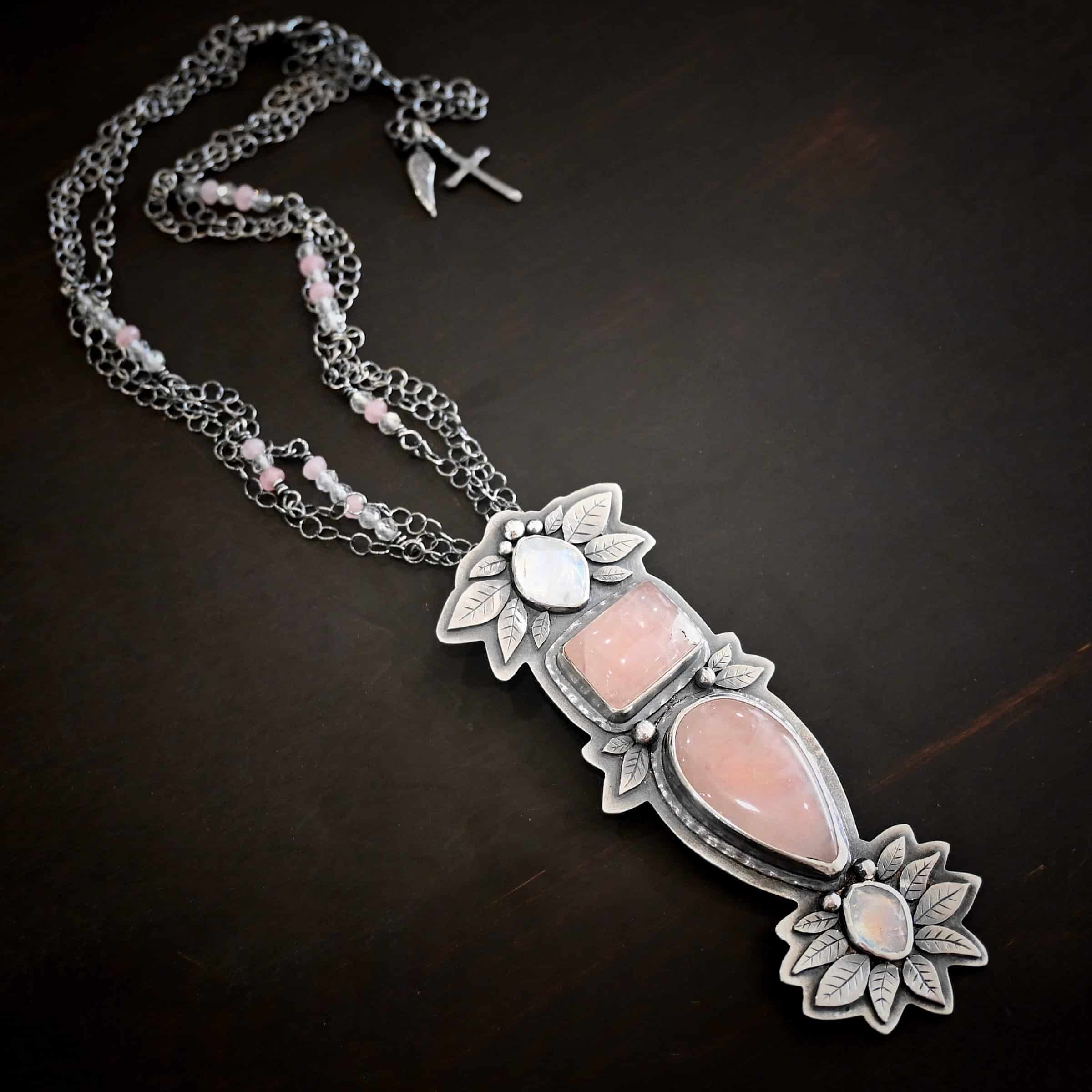 The Sparkling Lady Necklace Close up of the "Sparkling Lady" Necklace. Made out of sterling silver, rose quartz, and moonstone. There are two rose quartz and two moonstone. The top of the pendant has an oval moonstone, with a square rose quartz beneath it. Beneath that is another rose quartz that is a teardrop shape with the point facing downwards. At the bottom of the pendant is another moonstone that is an oval shape. Surrounding each of these stones and around the entire pendant are silver leaves and dots. Pendant is laying against a dark background.