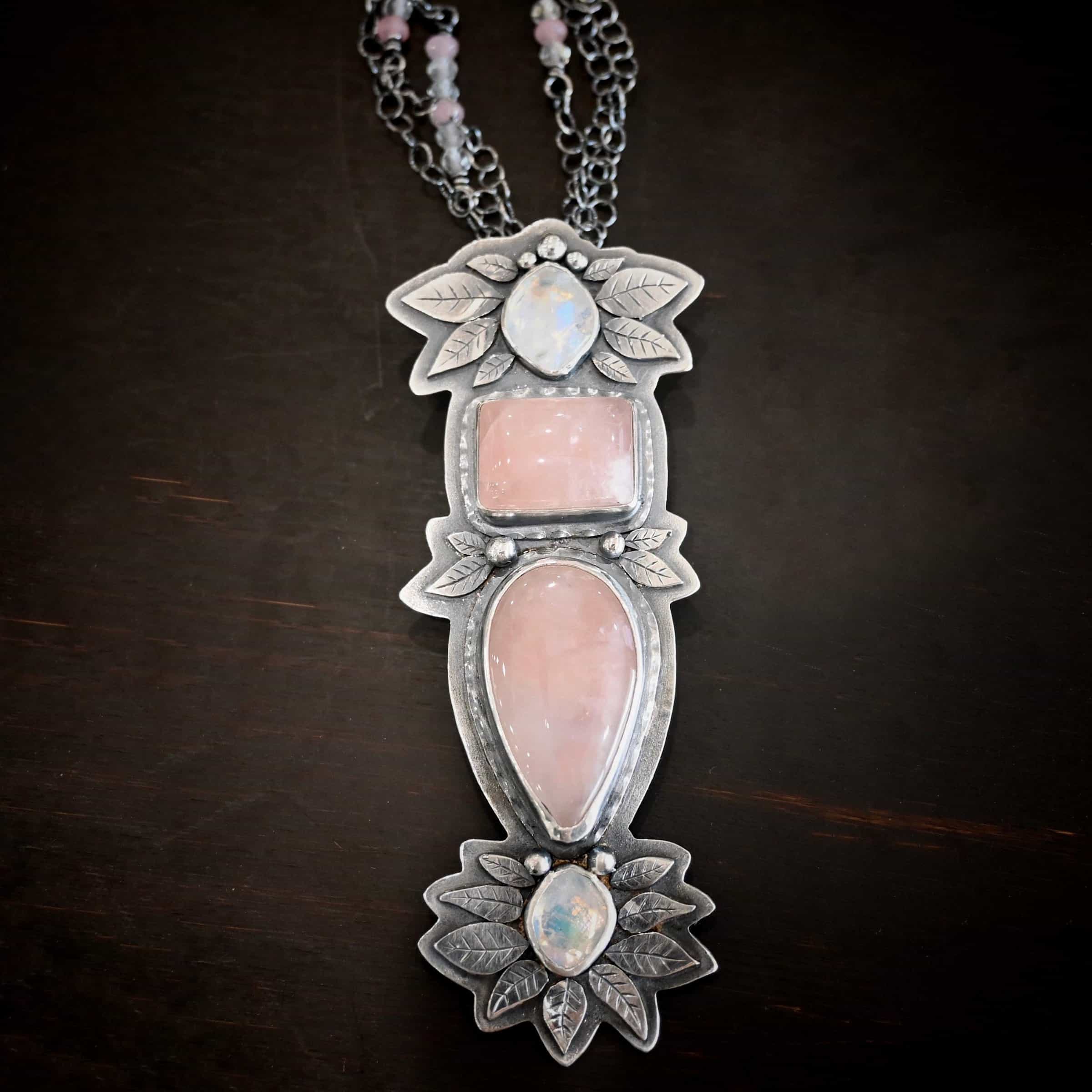 The Sparkling Lady Necklace Close up of the "Sparkling Lady" Necklace. Made out of sterling silver, rose quartz, and moonstone. There are two rose quartz and two moonstone. The top of the pendant has an oval moonstone, with a square rose quartz beneath it. Beneath that is another rose quartz that is a teardrop shape with the point facing downwards. At the bottom of the pendant is another moonstone that is an oval shape. Surrounding each of these stones and around the entire pendant are silver leaves and dots. Pendant is laying against a dark background.