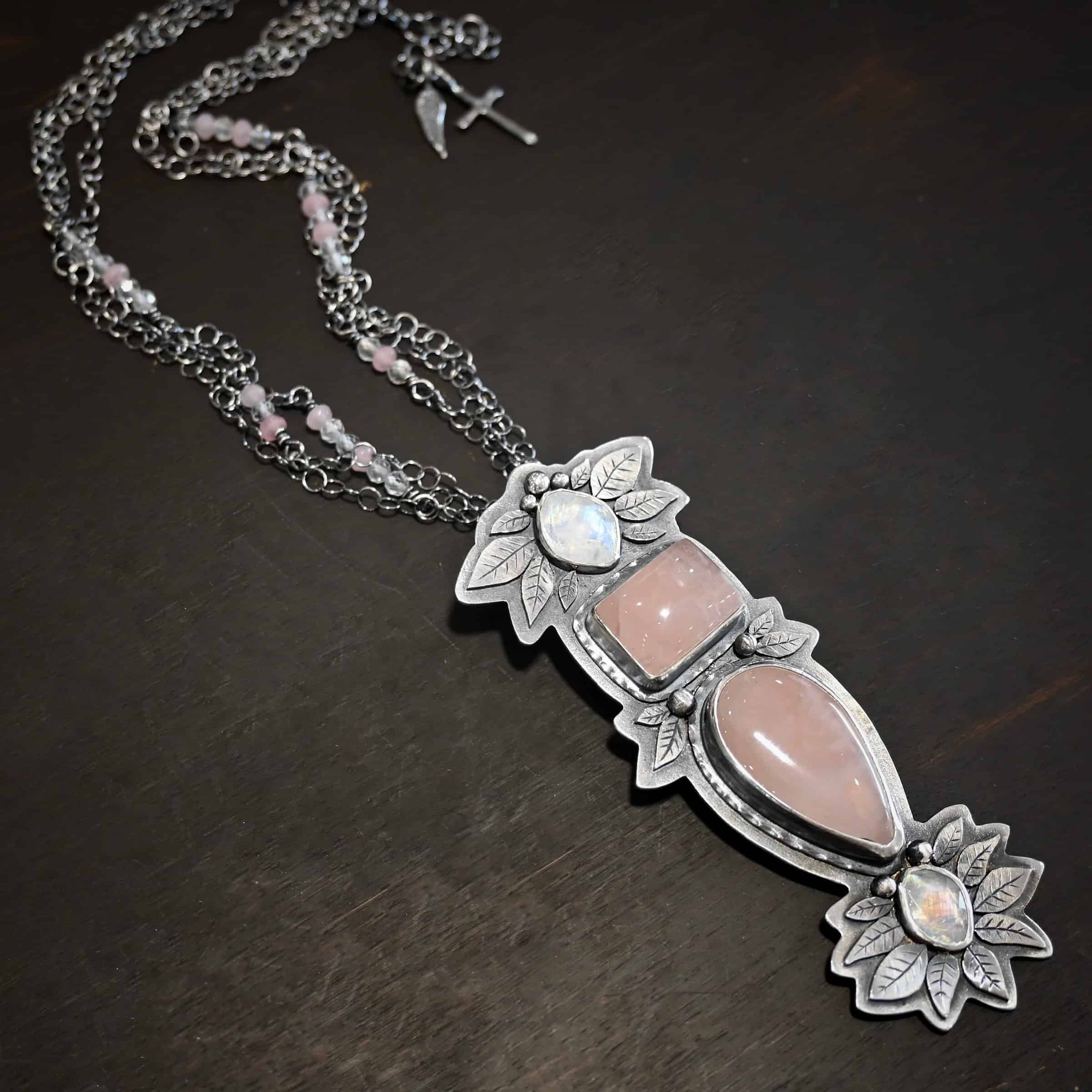 The Sparkling Lady Necklace Close up of the "Sparkling Lady" Necklace. Made out of sterling silver, rose quartz, and moonstone. There are two rose quartz and two moonstone. The top of the pendant has an oval moonstone, with a square rose quartz beneath it. Beneath that is another rose quartz that is a teardrop shape with the point facing downwards. At the bottom of the pendant is another moonstone that is an oval shape. Surrounding each of these stones and around the entire pendant are silver leaves and dots. Pendant is laying against a dark background.