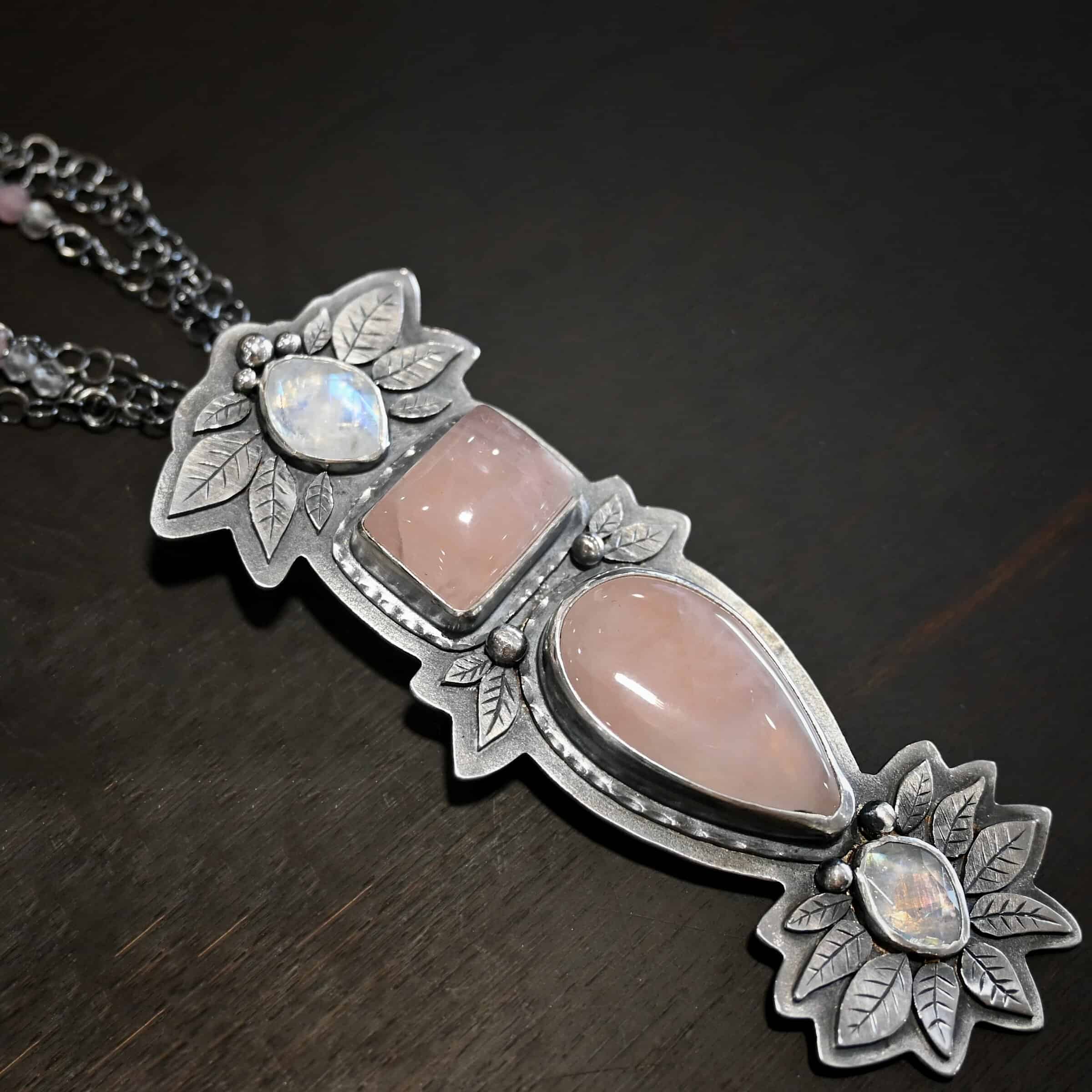 The Sparkling Lady Necklace Close up of the "Sparkling Lady" Necklace. Made out of sterling silver, rose quartz, and moonstone. There are two rose quartz and two moonstone. The top of the pendant has an oval moonstone, with a square rose quartz beneath it. Beneath that is another rose quartz that is a teardrop shape with the point facing downwards. At the bottom of the pendant is another moonstone that is an oval shape. Surrounding each of these stones and around the entire pendant are silver leaves and dots. Pendant is laying against a dark background.